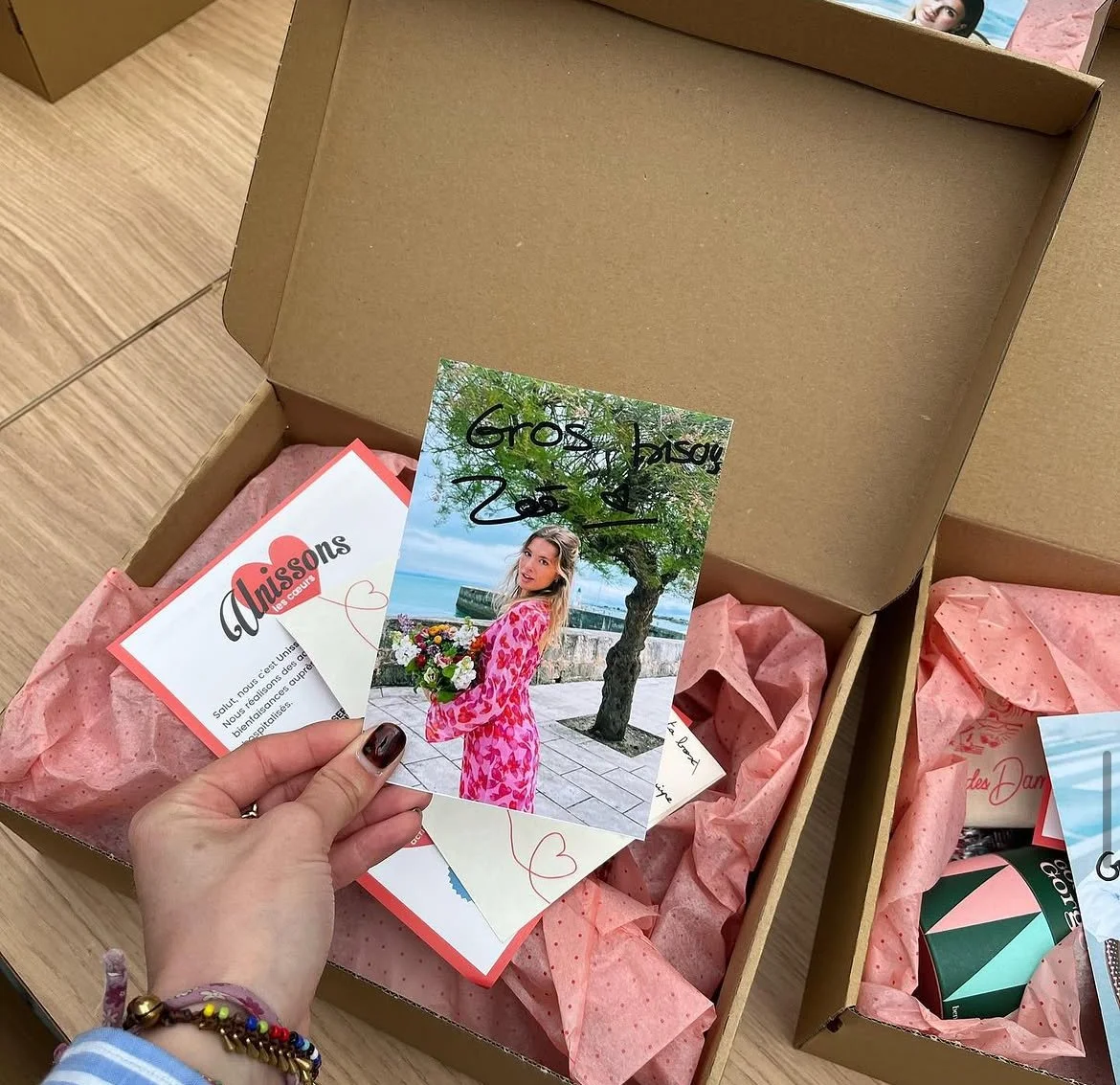 Une main tenant une carte photo d'une femme en robe rose à motifs floraux, dans une boîte en carton contenant du papier de soie rose et d'autres articles cadeaux.
