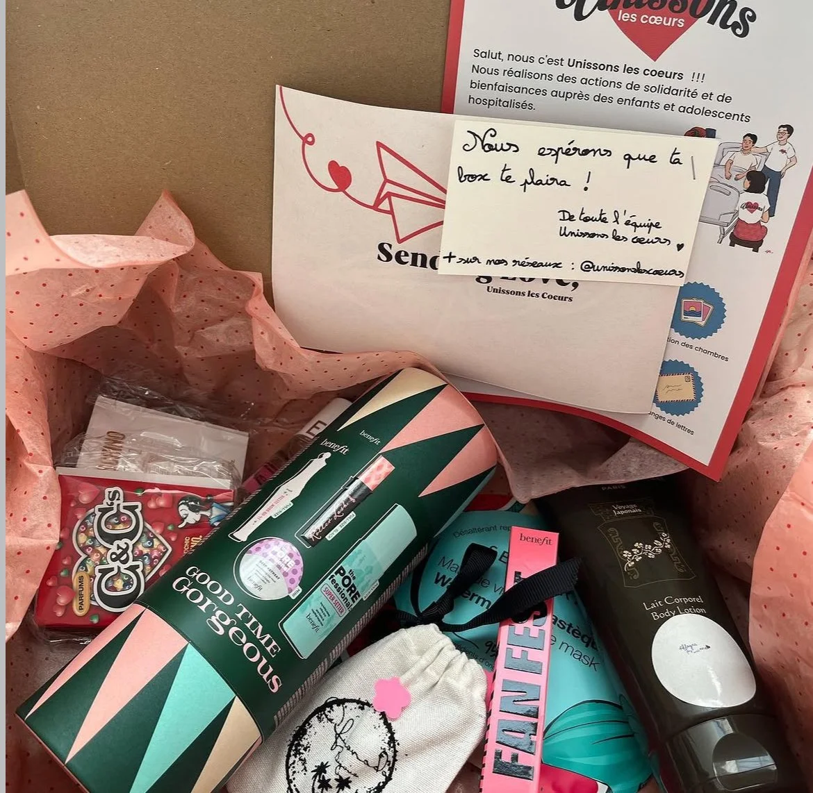 Cadeaux emballés avec un papier rose à pois rouges, comprenant divers produits de beauté, une boîte de bonbons, un sac en tissu avec un motif, et une carte avec un message manuscrit. Le tout présenté dans une boîte ou un panier.