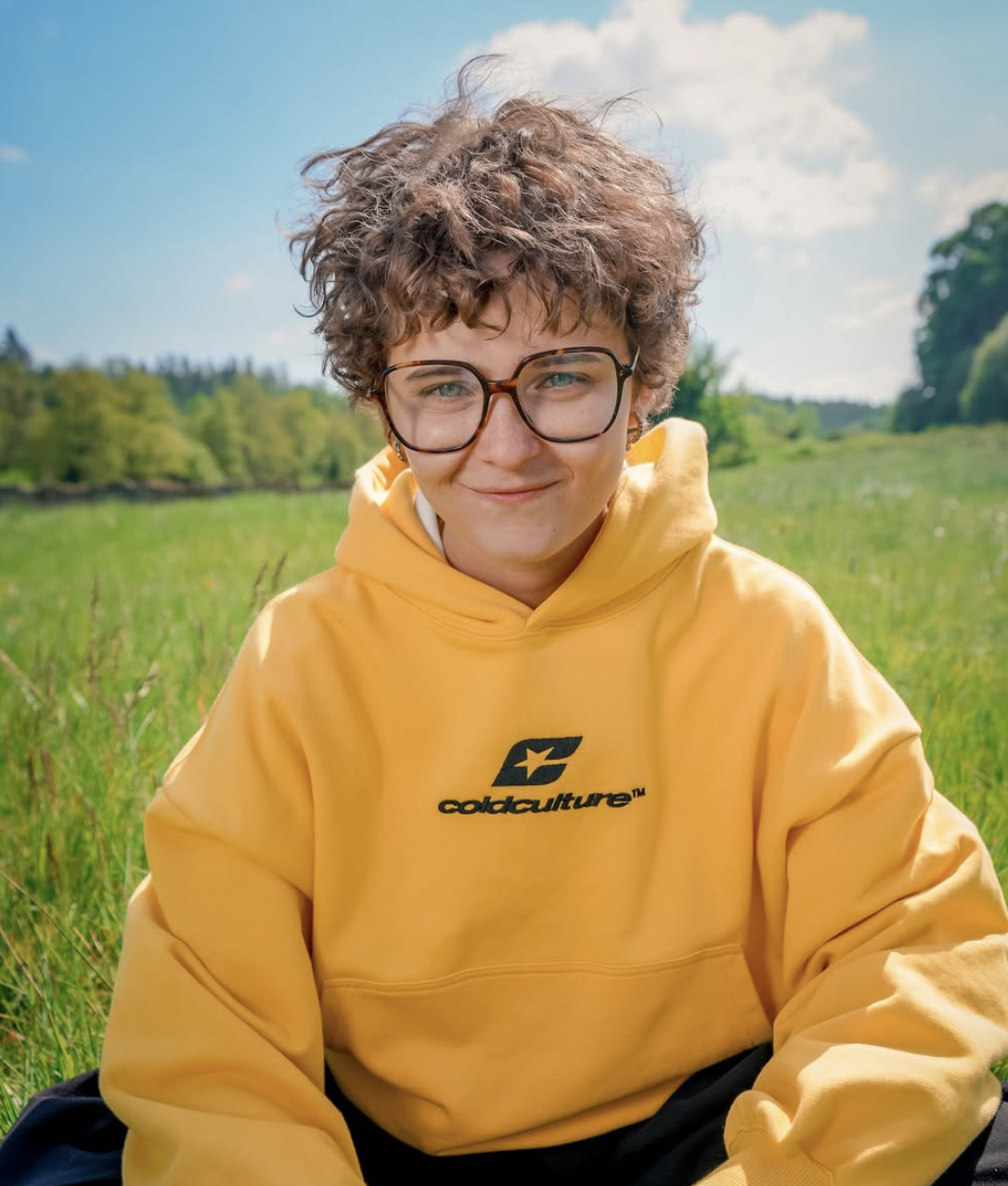 Un jeune homme avec des lunettes, portant un sweat à capuche jaune avec le logo 'coldculture', souriant dans un champ vert sous un ciel partiellement nuageux.