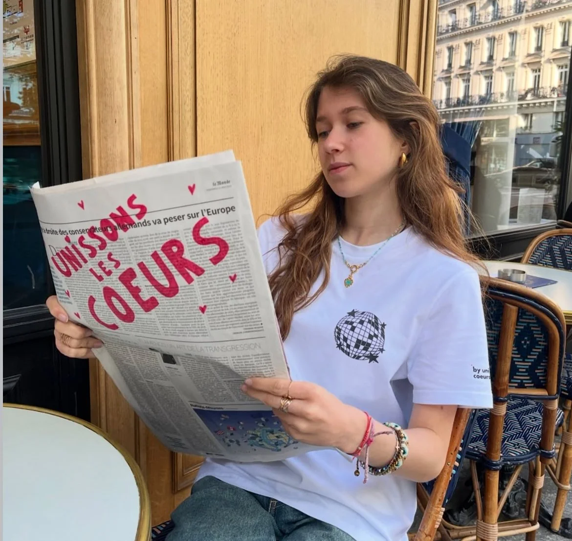 Jeune femme à lecture d'un journal dans un café parisien, portant un t-shirt blanc avec un logo globe et des bracelets colorés.