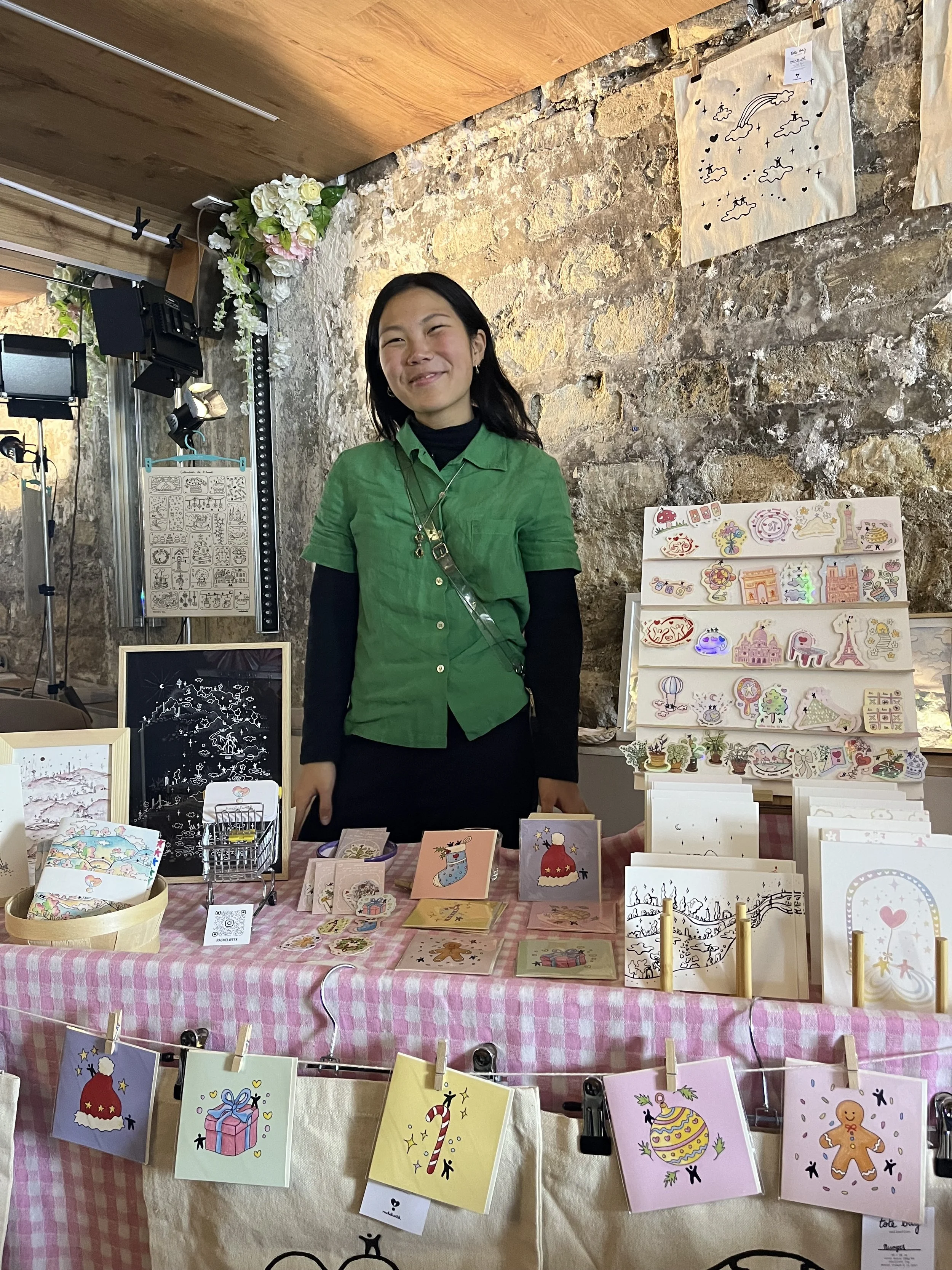 Femme souriante derrière un stand de souvenirs avec des cartes et accessoires aux motifs de Noël, décor en pierre et dessins sur le mur en arrière-plan.