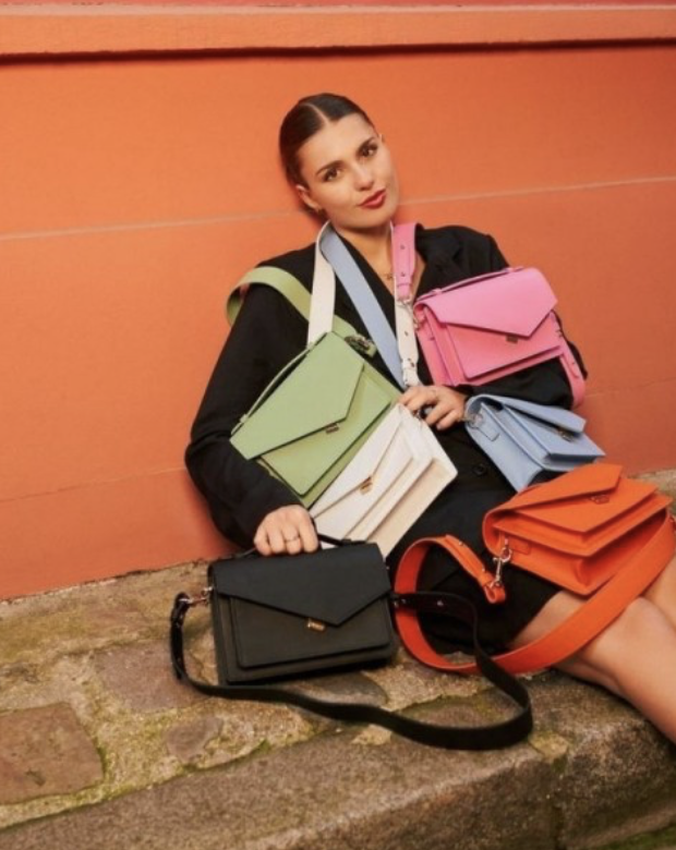 Une femme assise contre un mur orange portant plusieurs sacs à main de différentes couleurs, dont du vert, du blanc, du rose, du bleu, du noir et de l'orange.