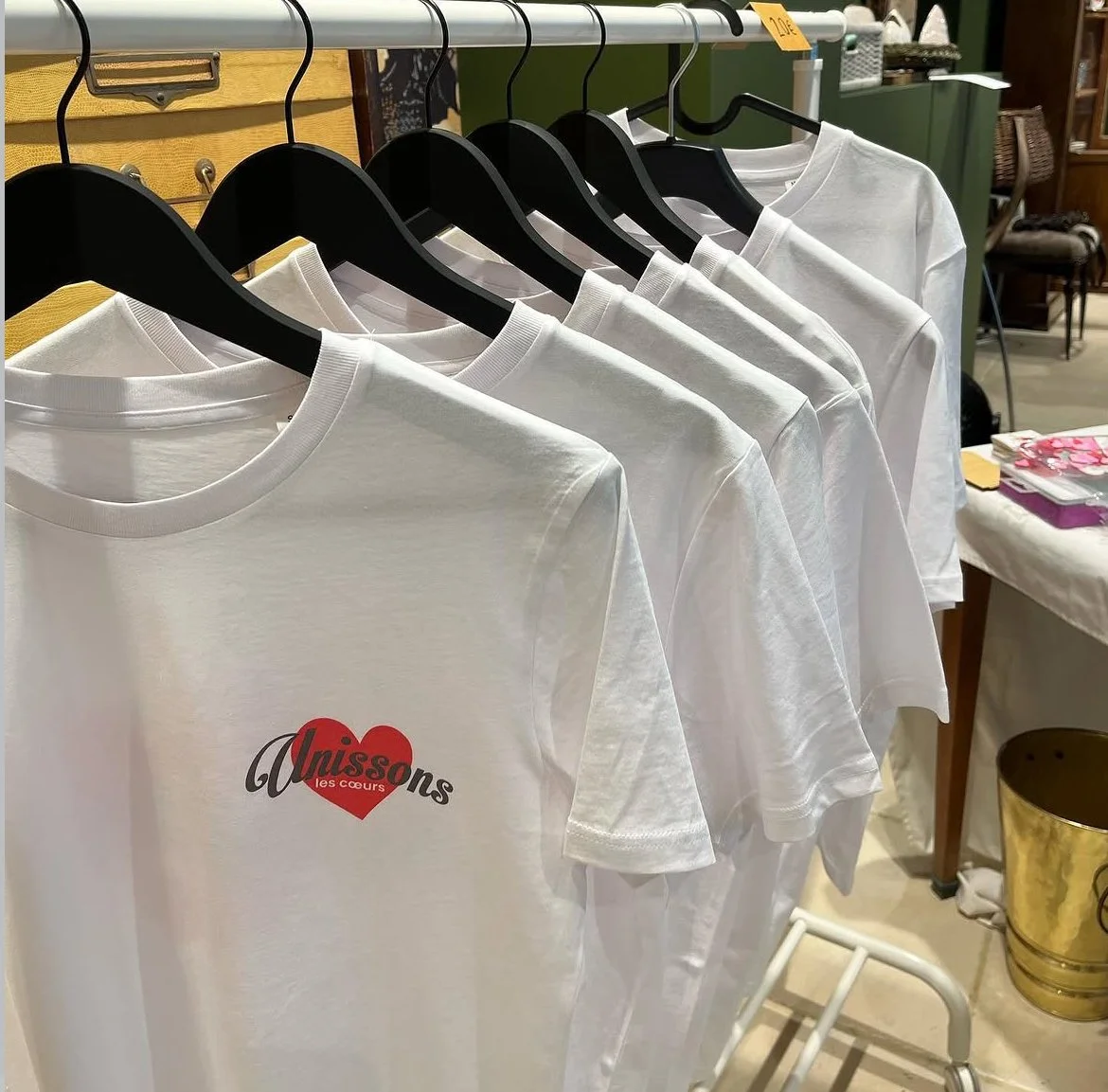 Une rangée de t-shirts blancs sur des cintres, avec un logo rouge et noir sur le devant indiquant "Auissones les cœurs".