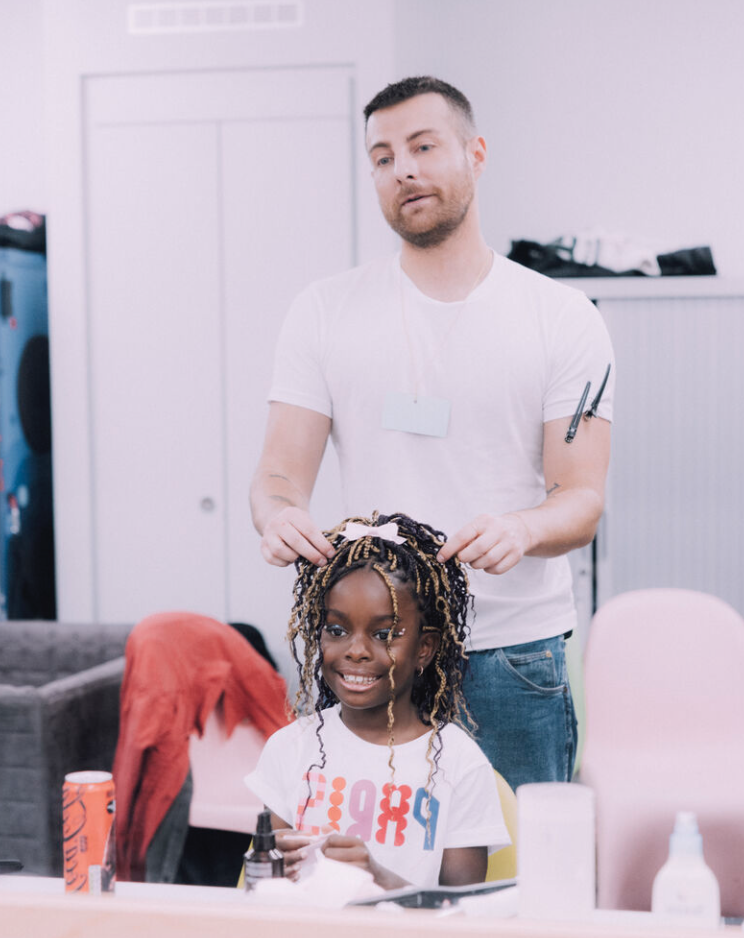 Un homme coiffe une petite fille aux cheveux bouclés et teints, dans un salon de coiffure, avec des produits capillaires sur la table.