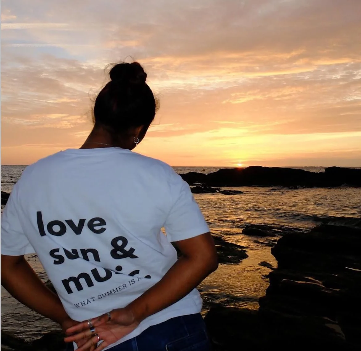 Une personne vue de dos regarde un coucher de soleil sur la mer, portant un t-shirt blanc avec l'inscription 'love sun & music'.