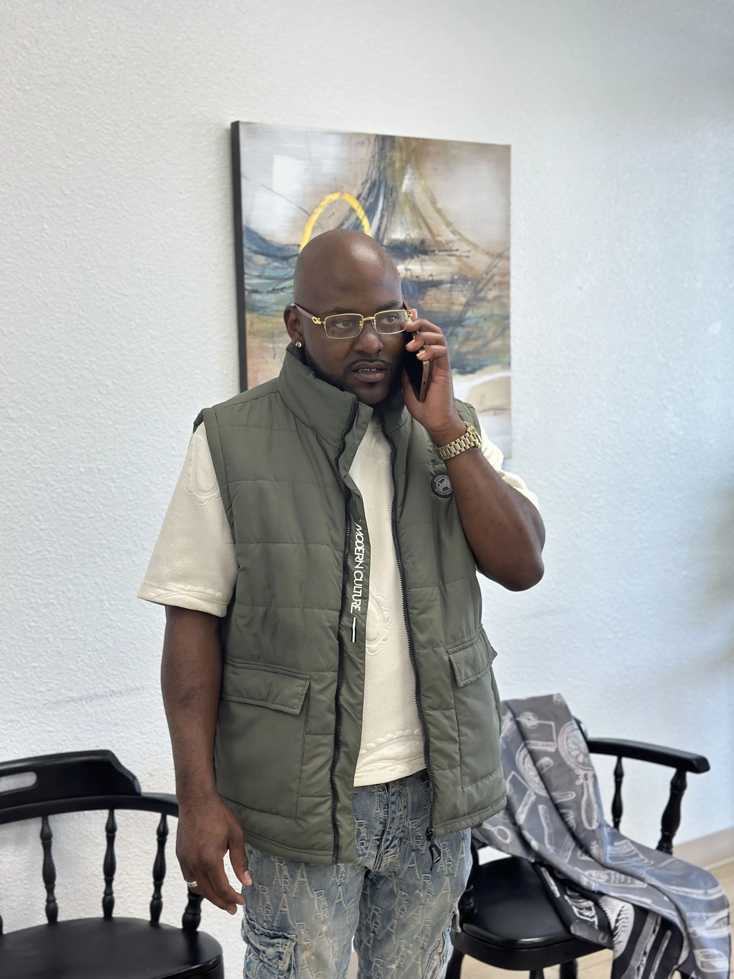 A bald man with glasses wearing a green vest and patterned jeans talking on a landline phone in a room with white walls. There are black chairs and a painting on the wall behind him, and a patterned blanket draped over the back of one of the chairs.