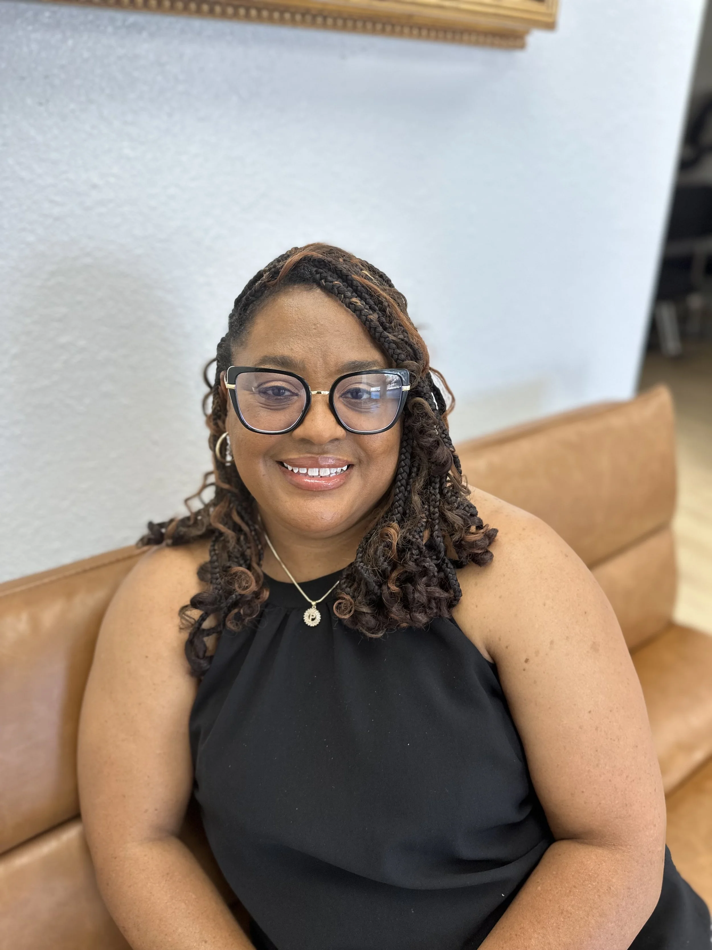 A smiling woman with braids and glasses wearing a black dress sitting on a tan leather couch in a room with a white wall.