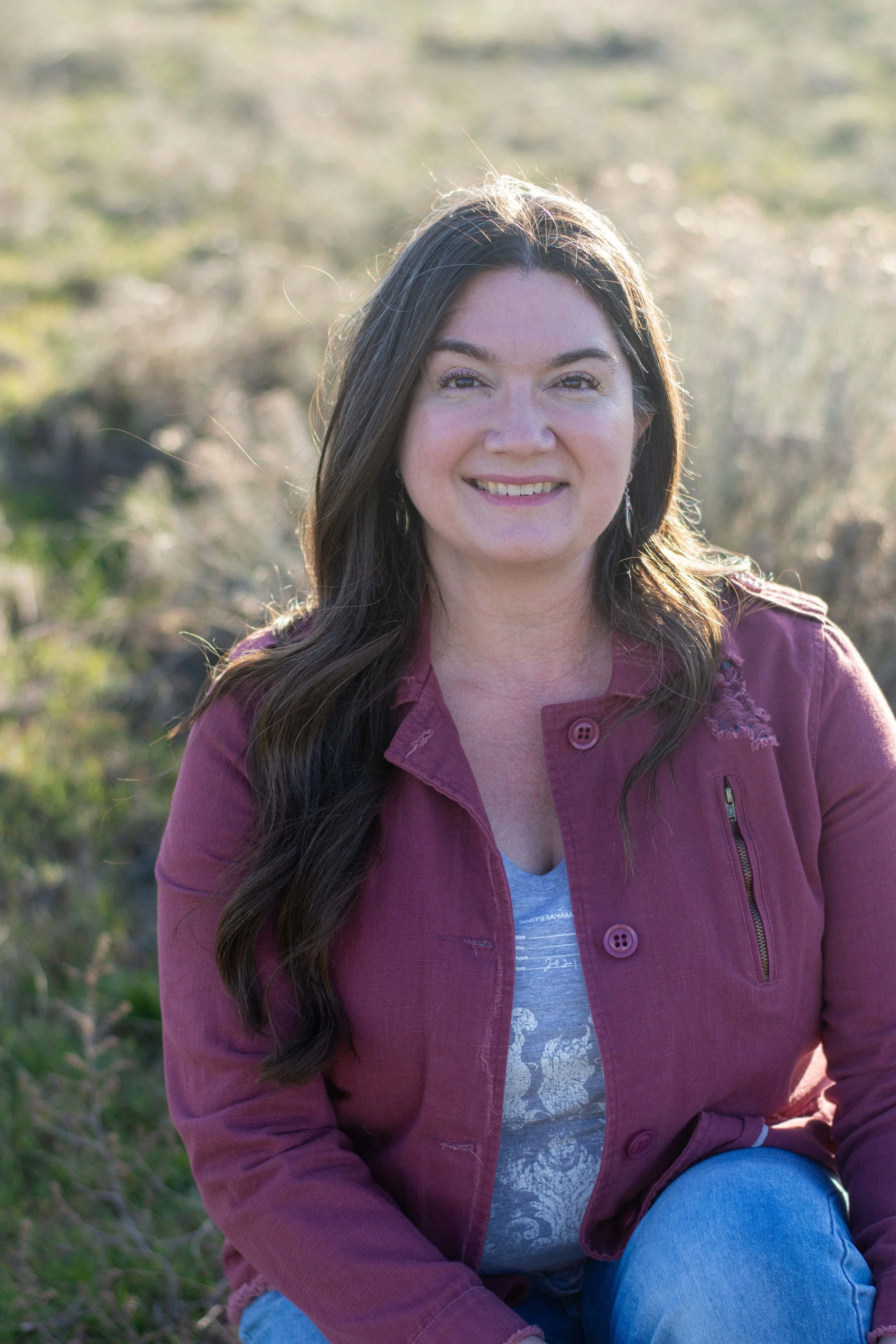 A woman with long dark hair wearing a maroon jacket and light blue jeans sitting outdoors on grass with a blurred background of greenery and sunlight.