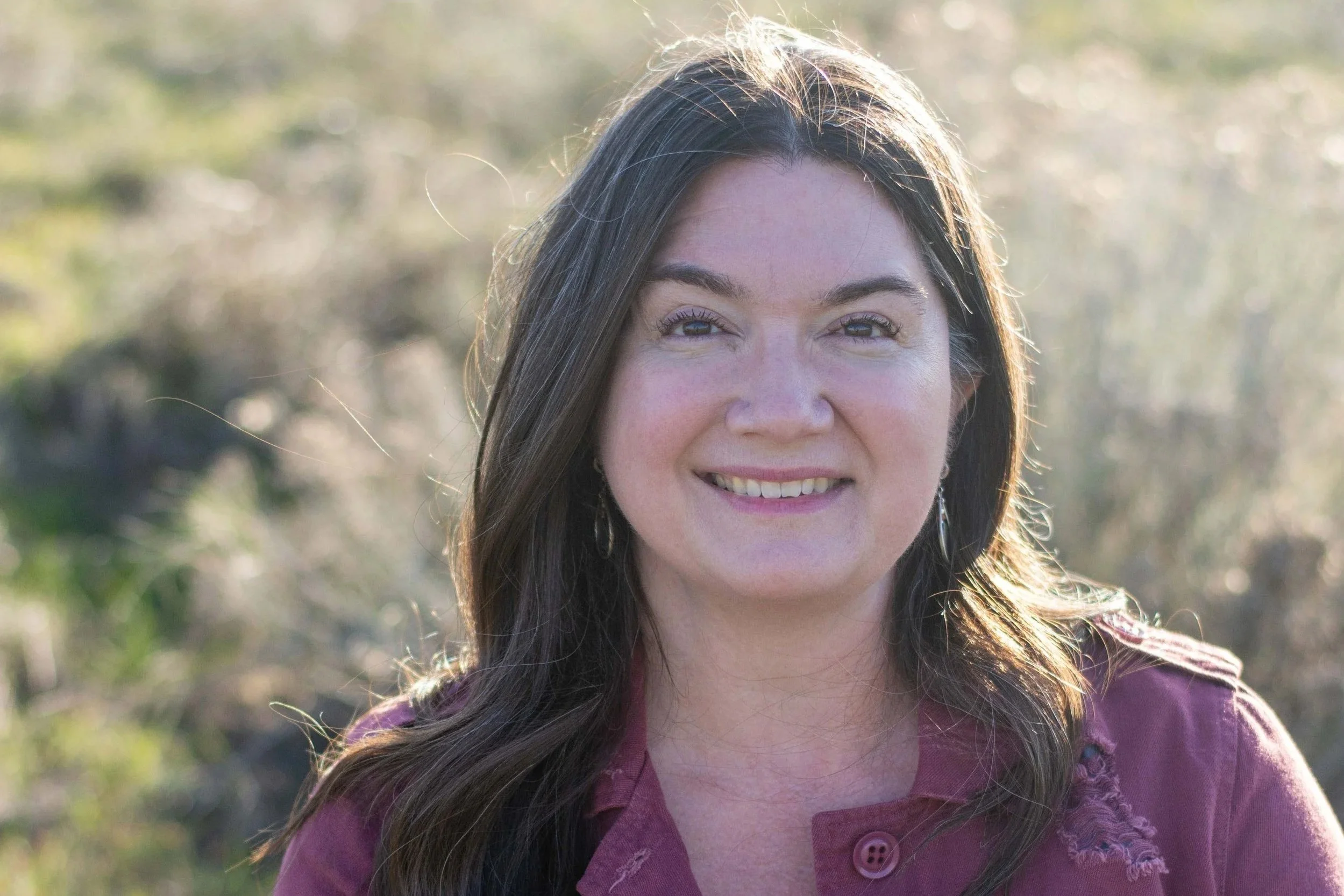 A woman with long brown hair smiling outdoors in natural sunlight, wearing a purple jacket.