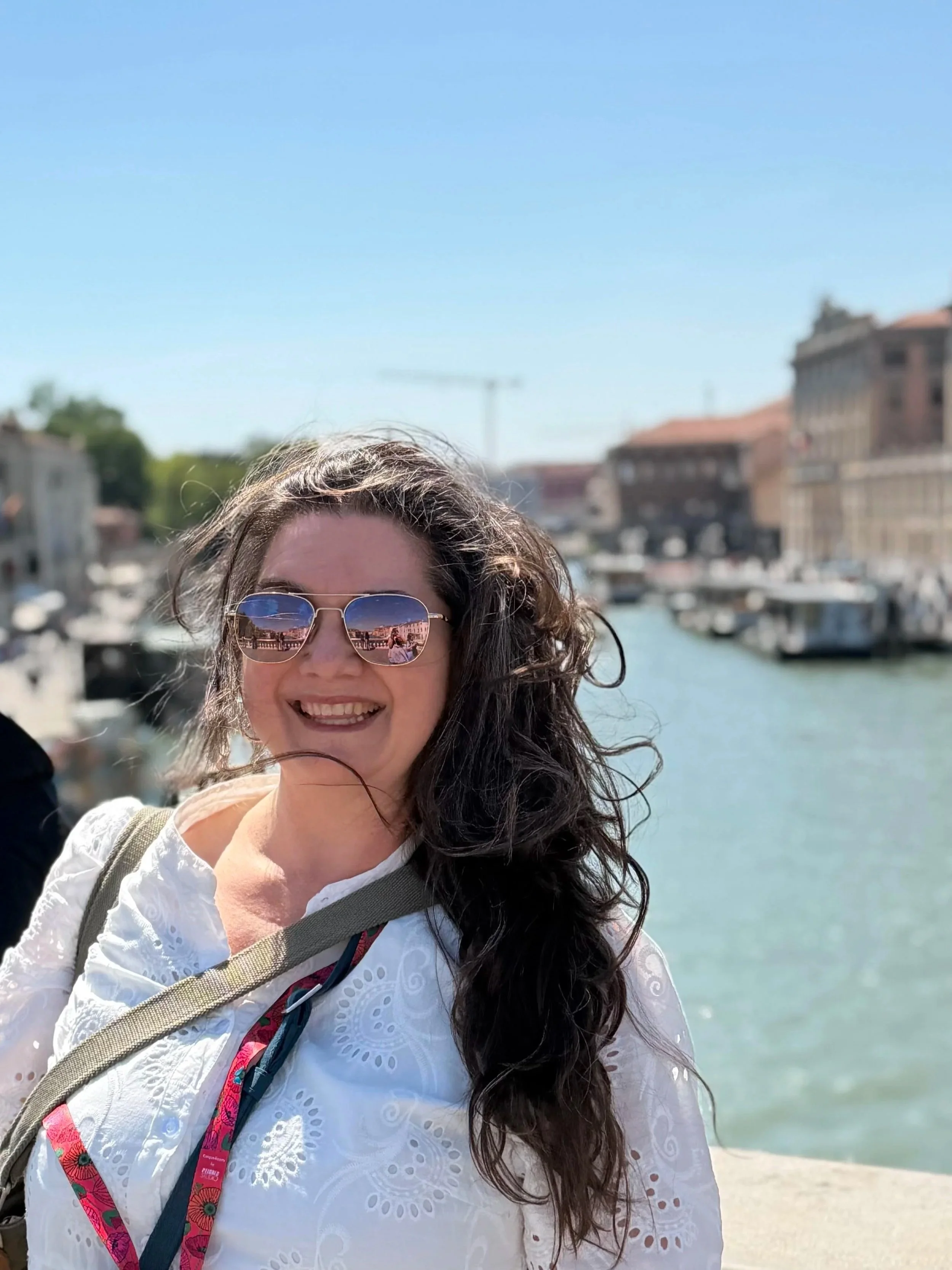 A woman with long curly dark hair wearing reflective sunglasses, a white embroidered blouse, and a beige strap bag, standing on a sunny beach with boats and buildings in the background.