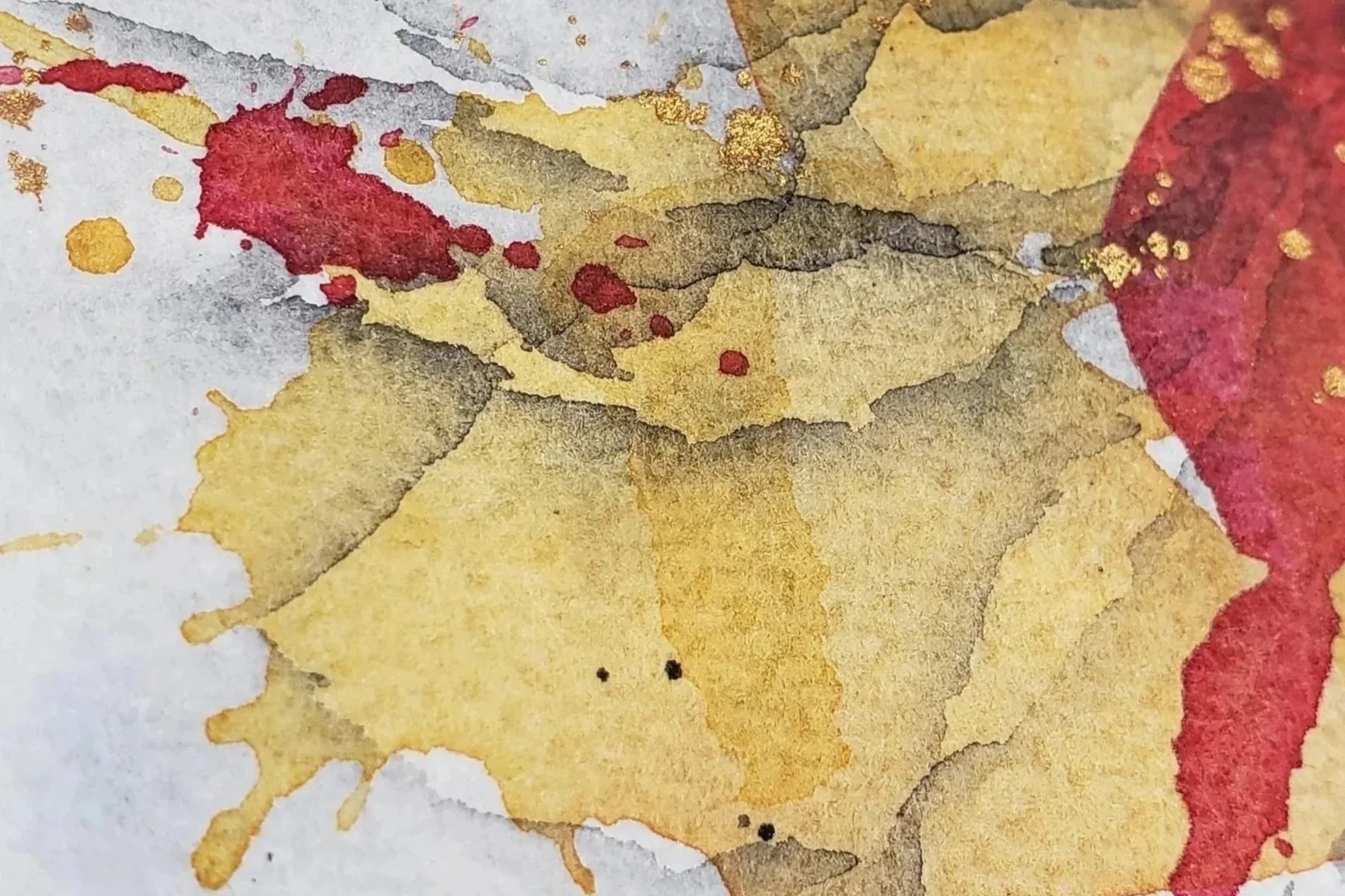 Close-up of watercolor paint splashes in shades of yellow, brown, red, and gray on paper.