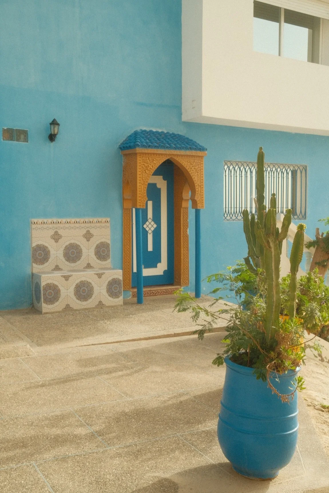 The image shows a blue exterior wall of a building with a Moroccan-style door featuring intricate designs and a small tiled bench. To the right, there is a tall cactus and other plants in a blue pot. The ground is tiled, and a small exterior light fi