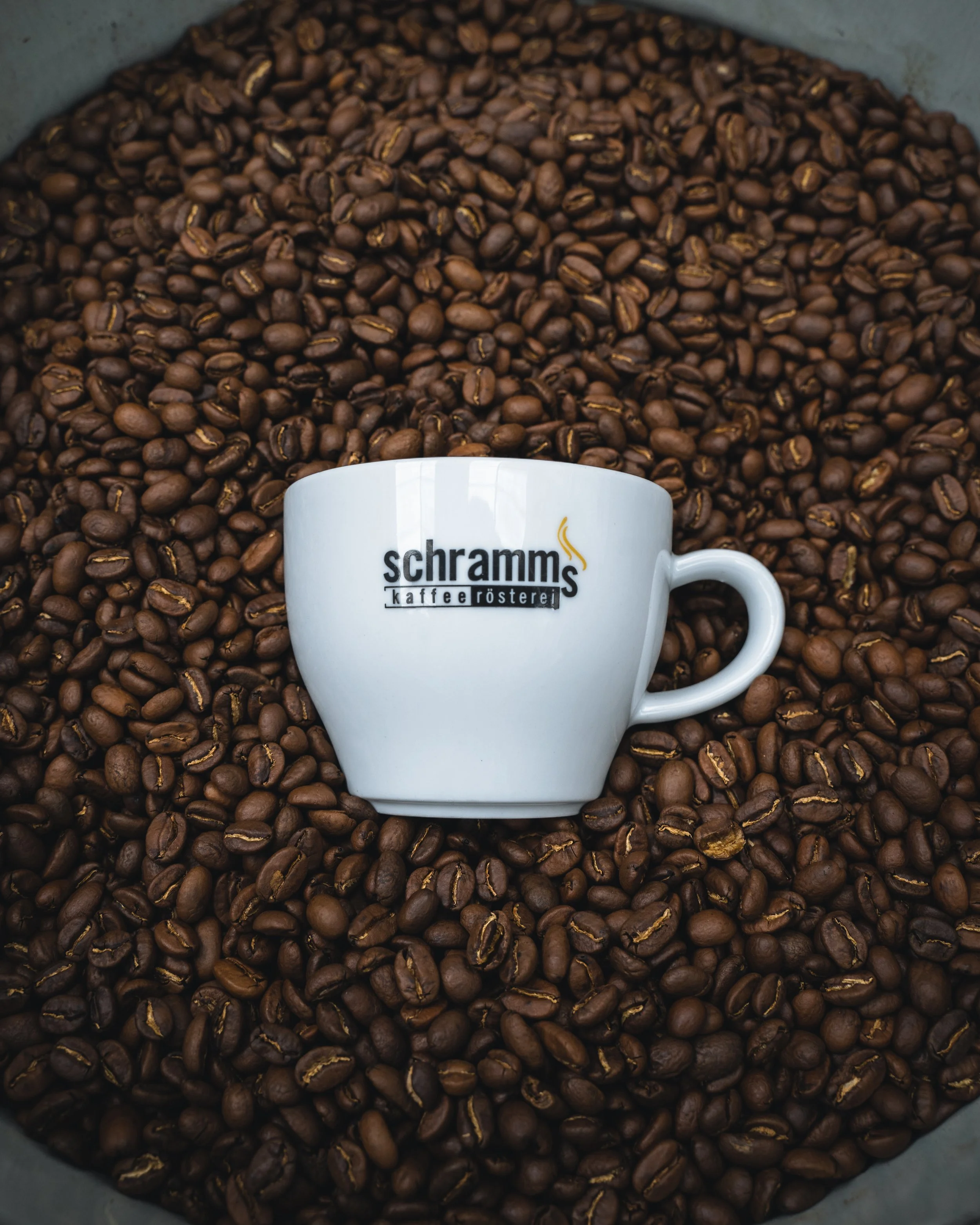 A white coffee mug with the logo 'schrammskaffee röster' sitting on top of a large pile of roasted coffee beans.