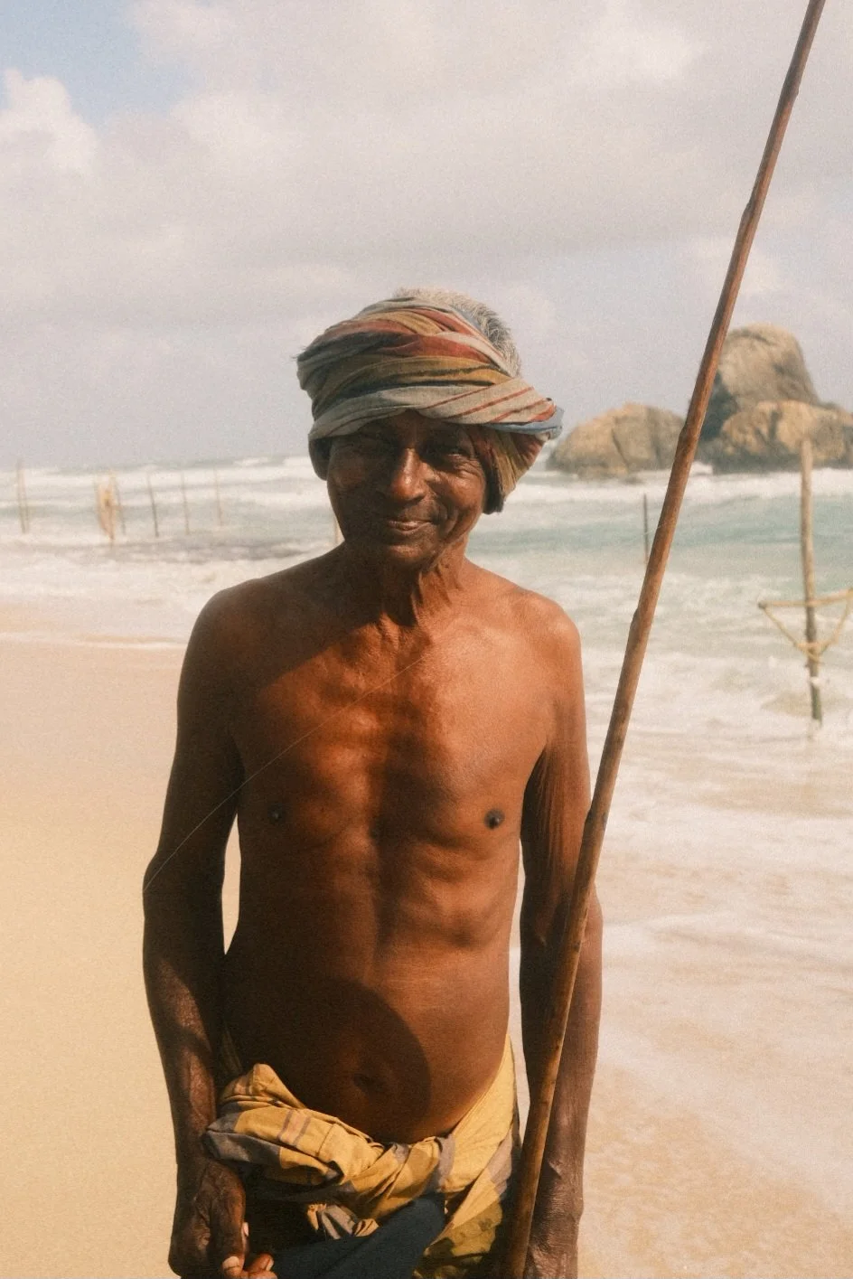 An elderly man with dark skin standing shirtless on a beach, wearing a colorful wrapped headscarf and holding a long wooden stick, with the ocean, rocks, and a cloudy sky in the background.