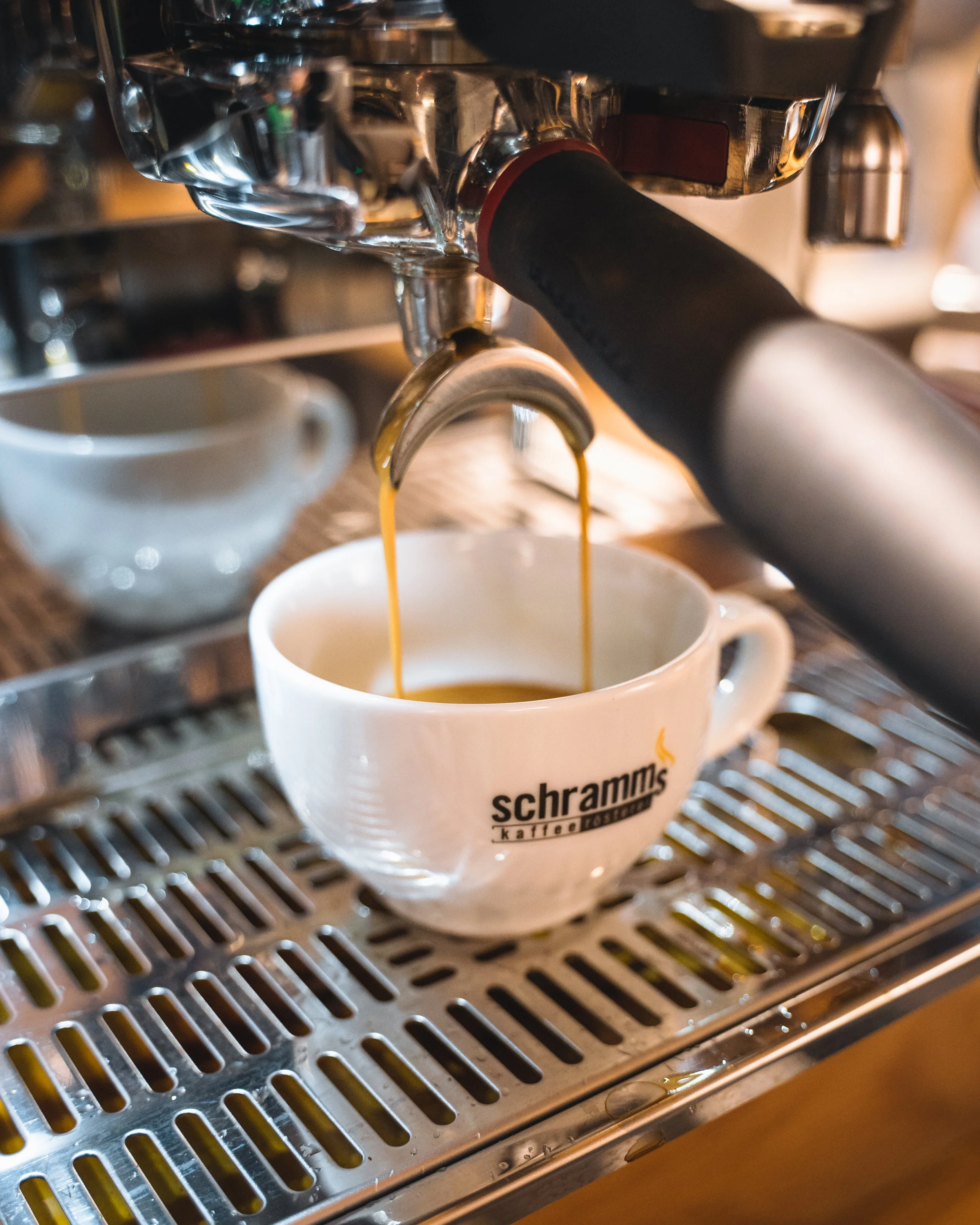Close-up view of coffee being brewed from an espresso machine into a white coffee cup with the logo 'schramm's kaffee' on it, on a stainless steel drip tray.