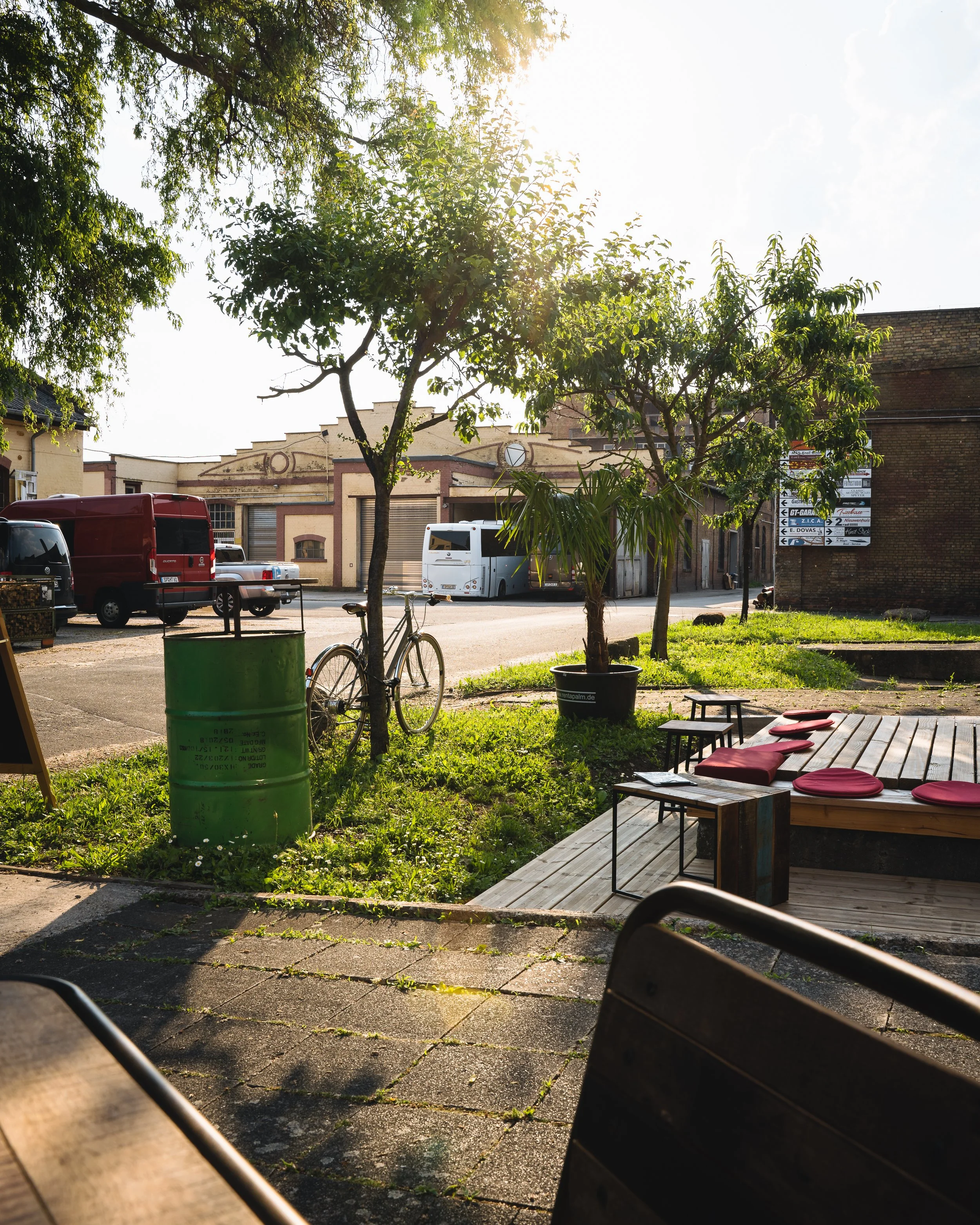 An outdoor scene showing a small patio area with a wooden platform, red cushions, and outdoor furniture. There are trees, parked vehicles, and a brick building with a signpost in the background. The sun is shining, creating a bright and warm atmosphe