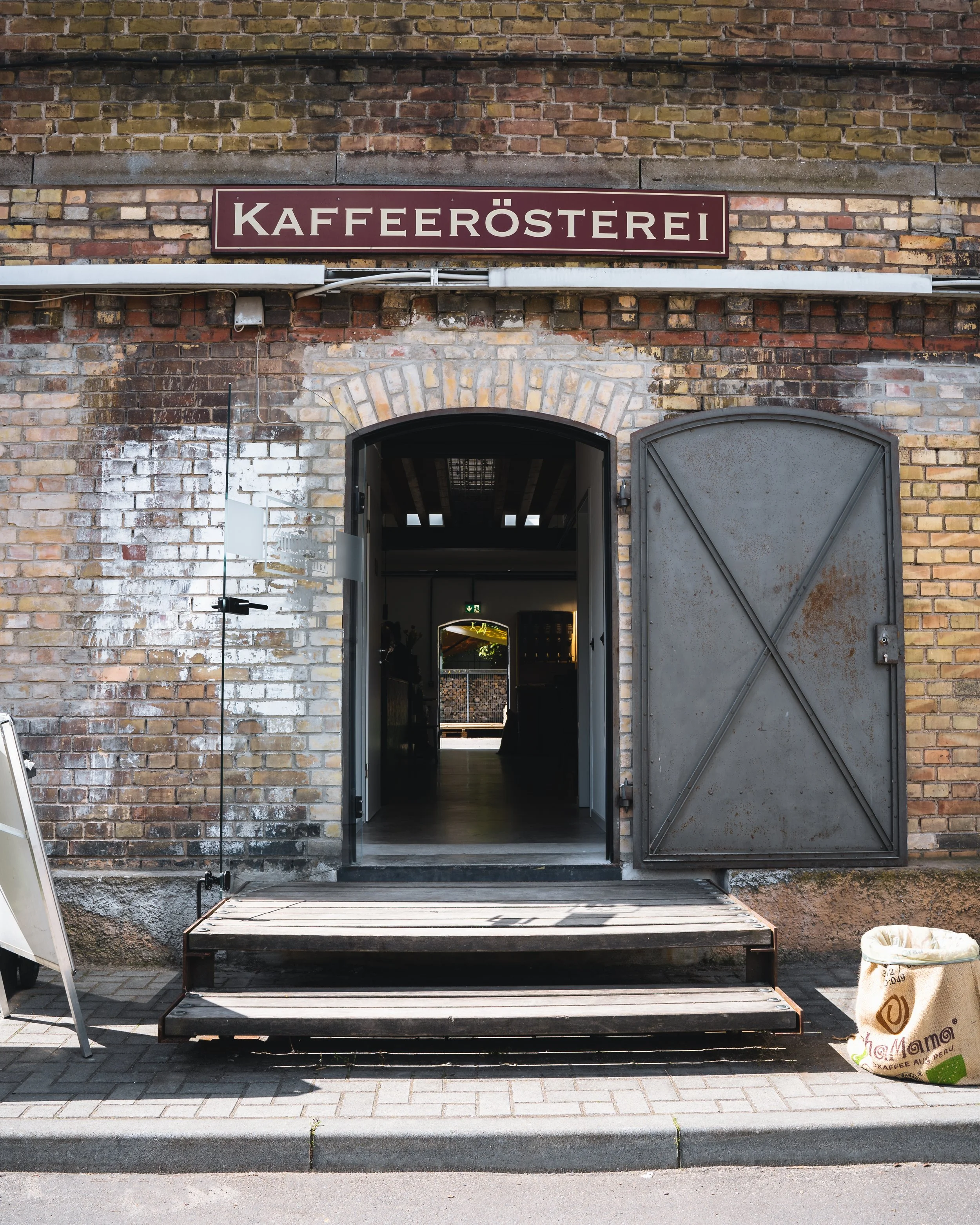 Brick building with a sign reading 'Kaffeerösterei' above an entrance, with a set of wooden stairs leading up to it and a large paper bag of coffee on the ground nearby.