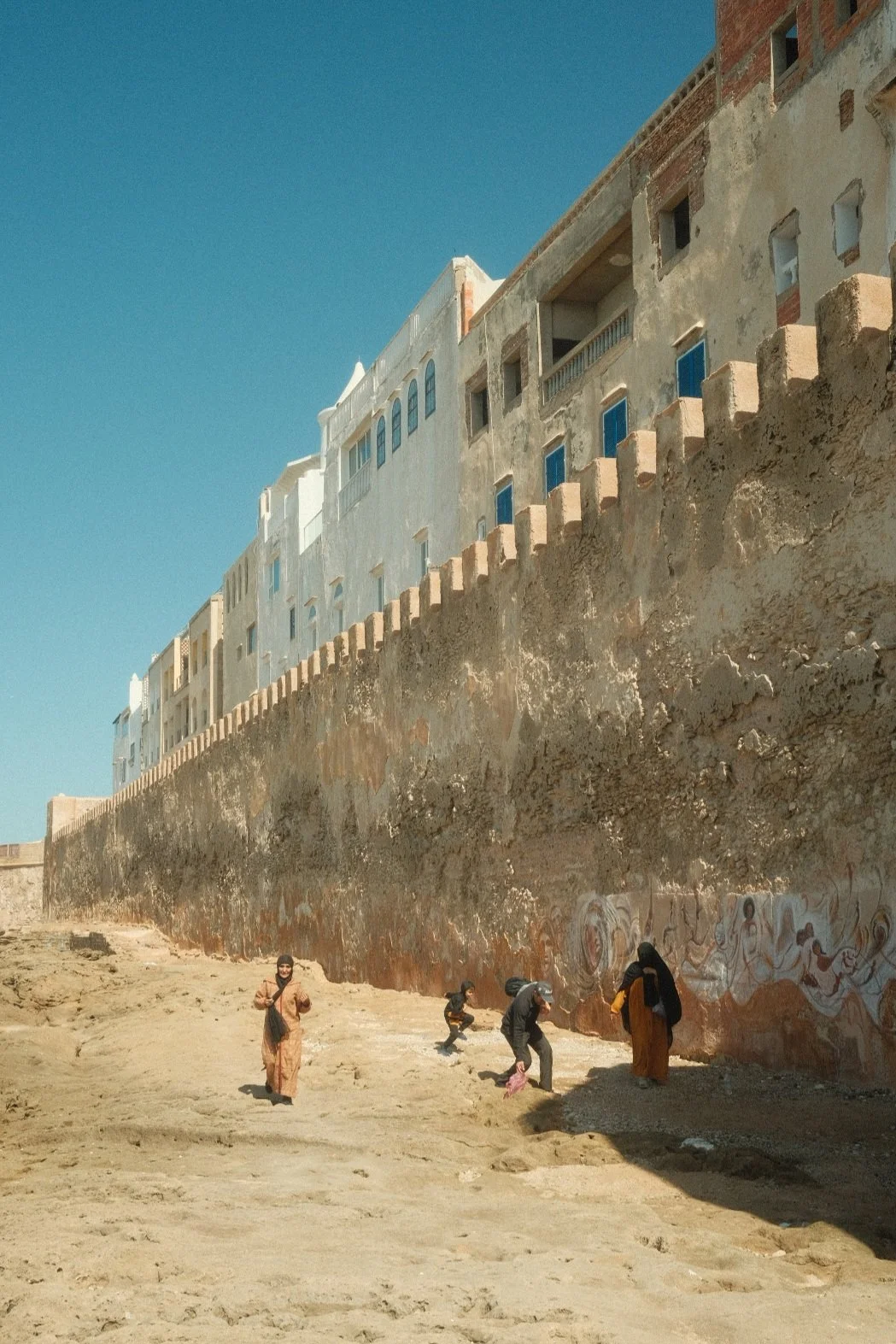 People walking and playing near a large, weathered wall with buildings on top against a clear blue sky.