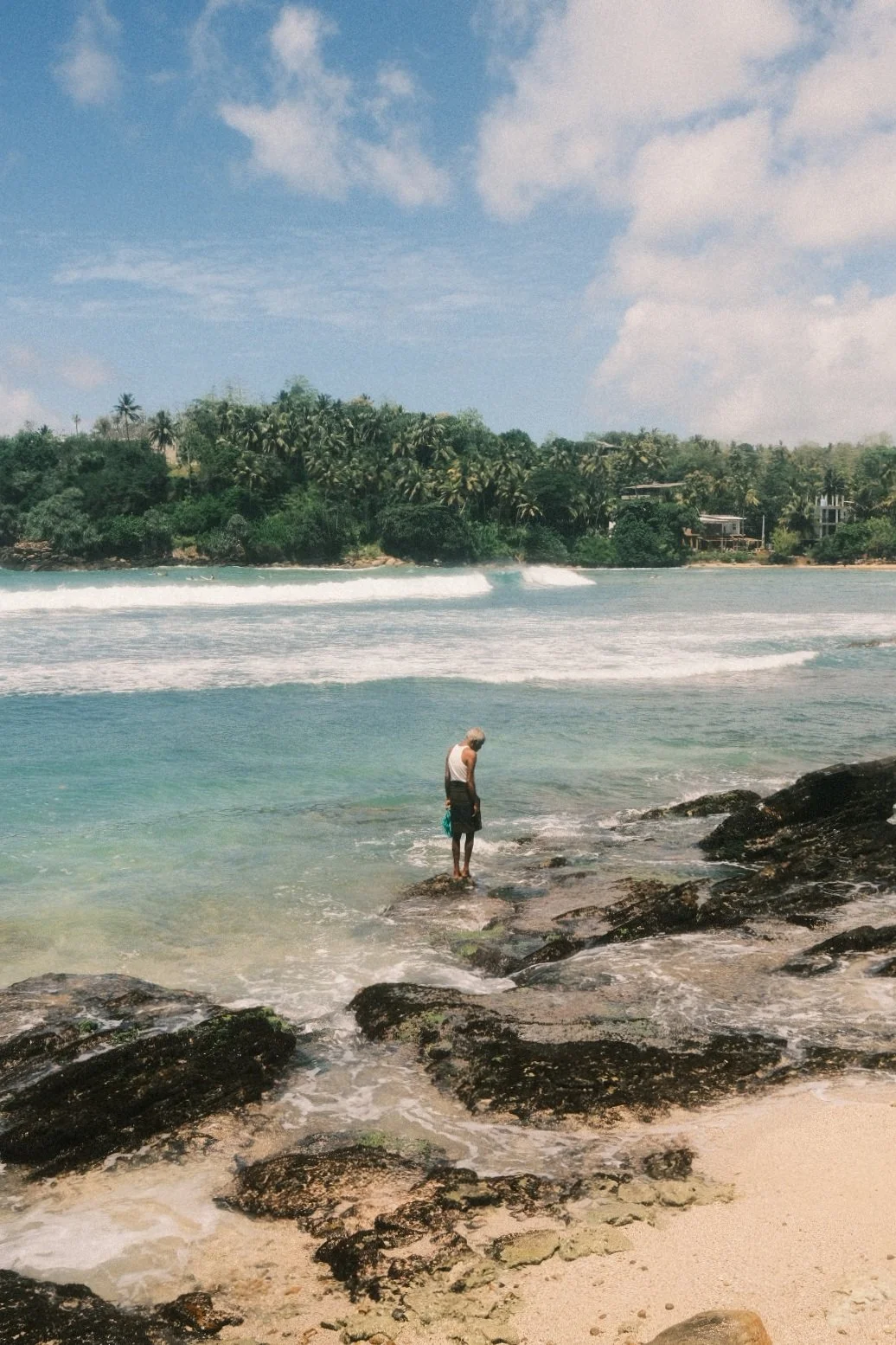A person standing on rocks at the edge of the ocean, looking down at the water, with a backdrop of lush green trees and buildings along the inland shoreline under a partly cloudy sky.