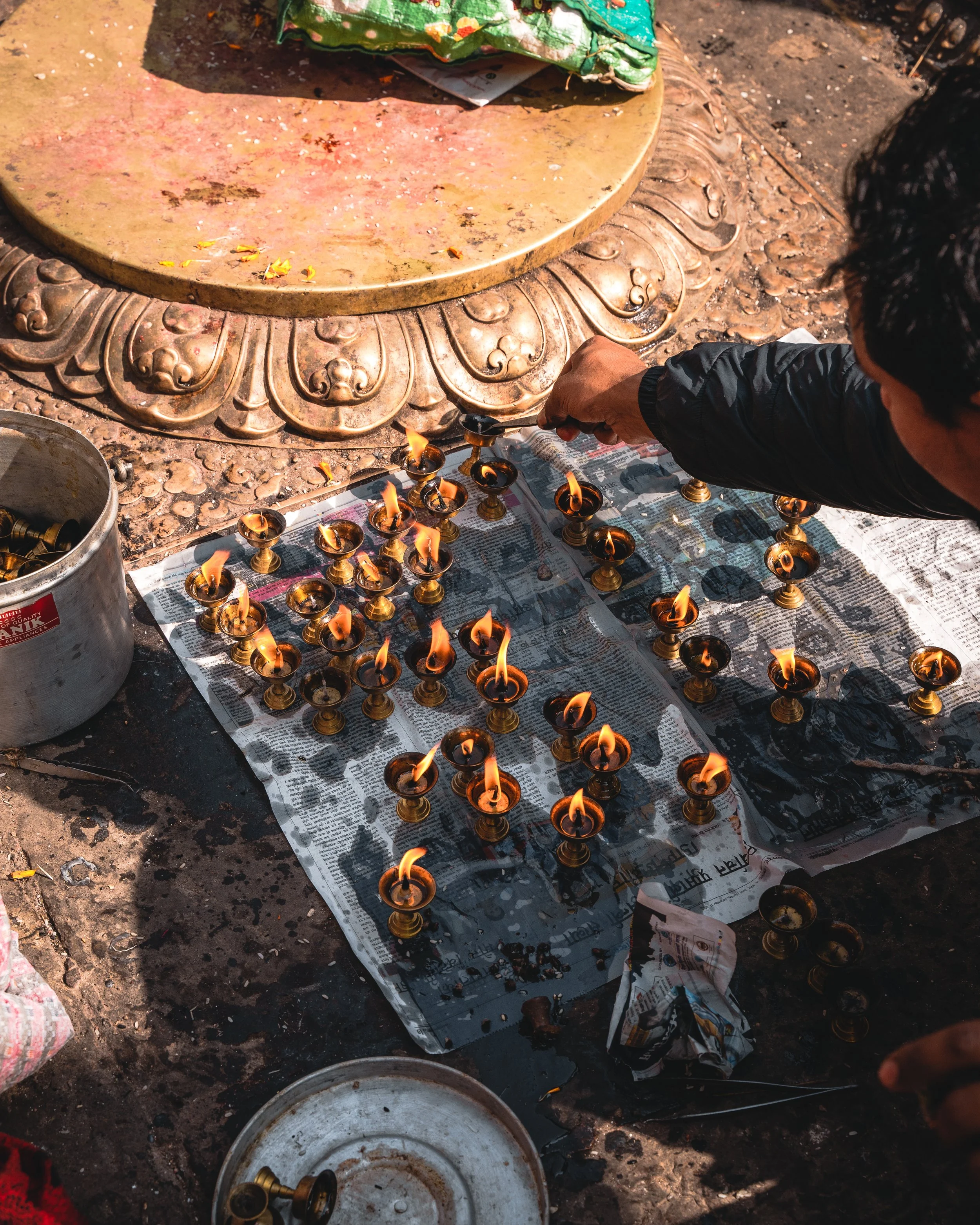 A person lighting small oil lamps placed on a newspaper-covered ground, with a large decorative metal plate in the background.