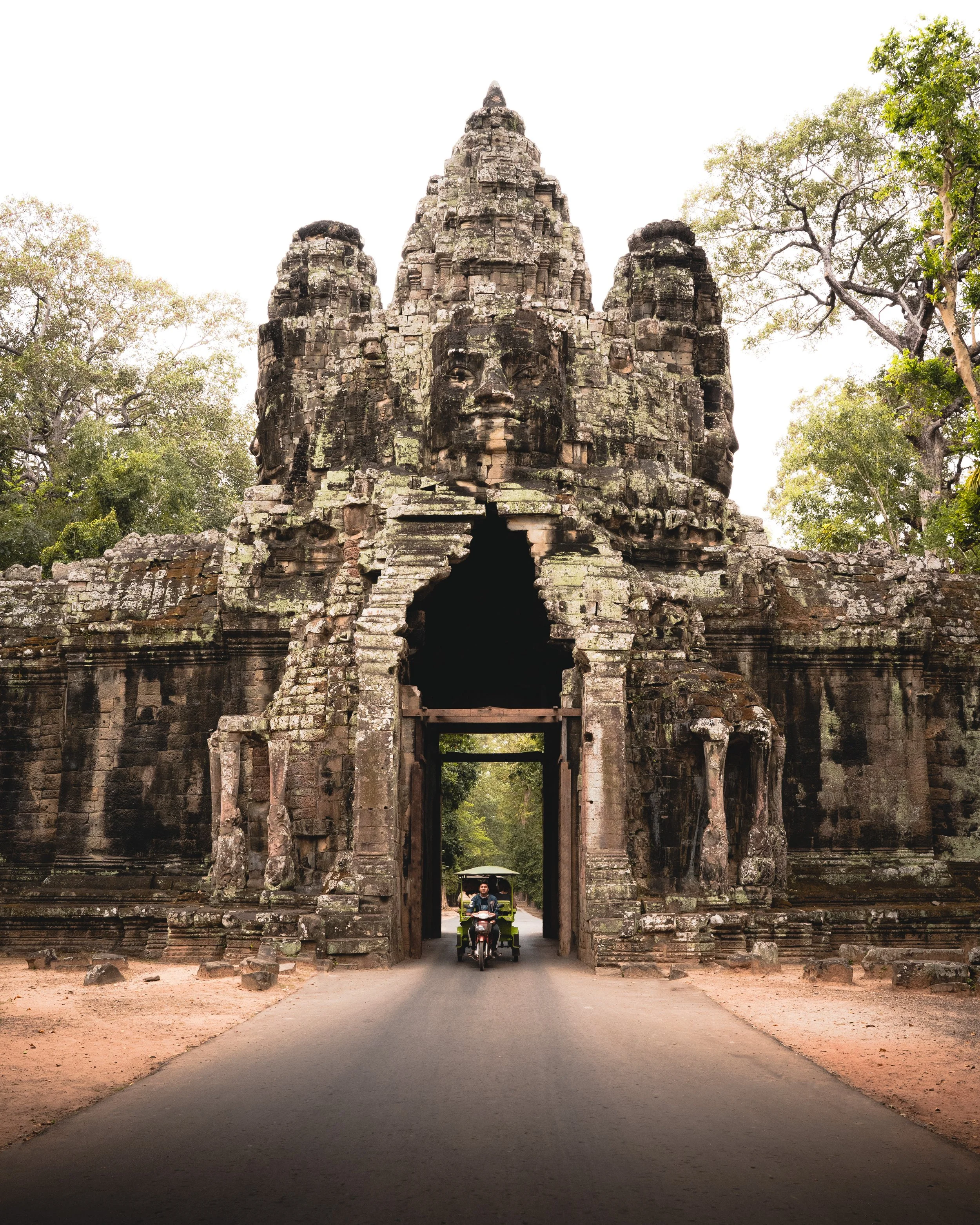 Ancient stone gateway with large carved faces, trees on either side, and a person riding a motorcycle with a sidecar passing through.