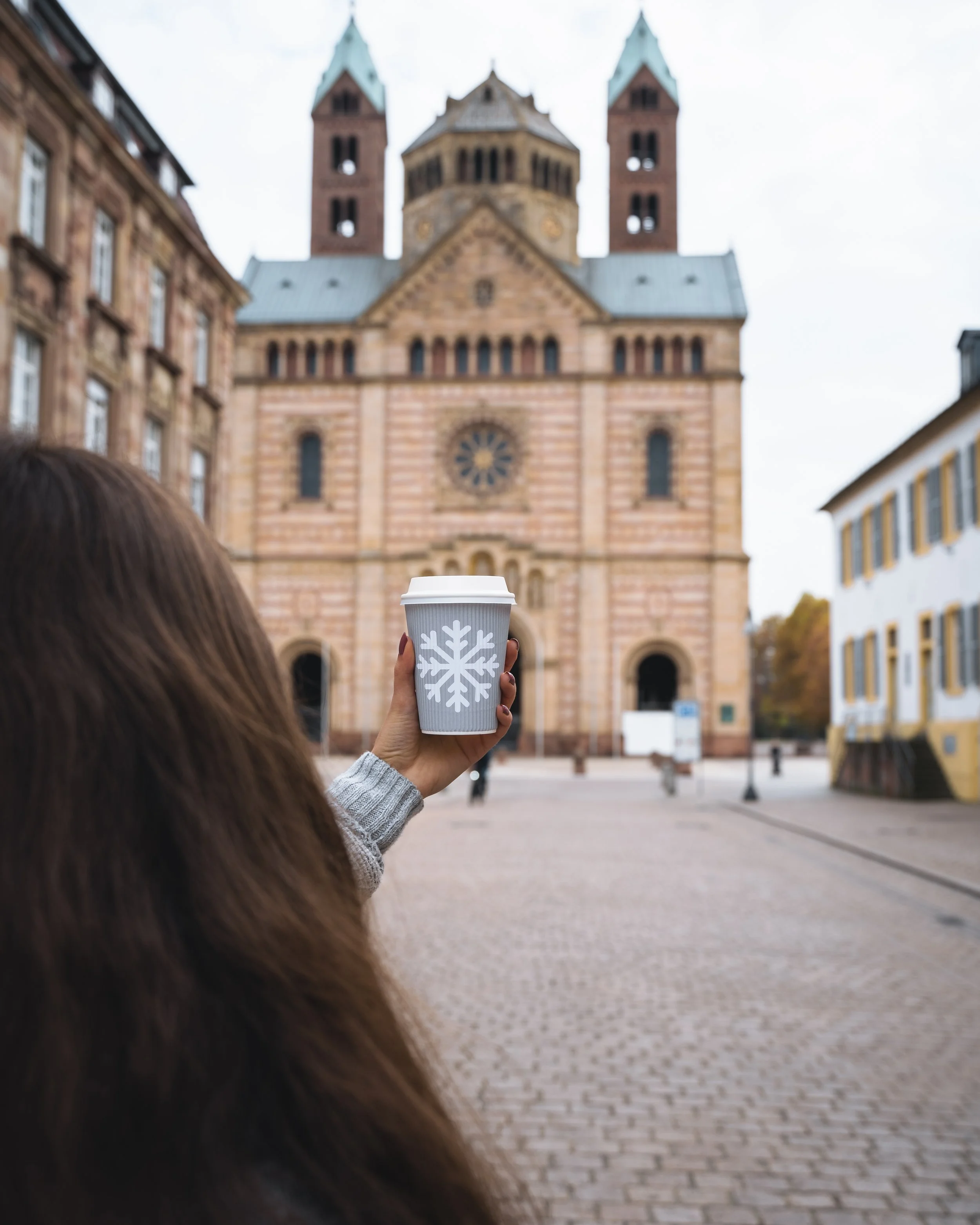 A person holding a to-go coffee cup with a snowflake design in front of the Frauenkirche church in Munich, Germany, with cobblestone street and adjacent buildings.