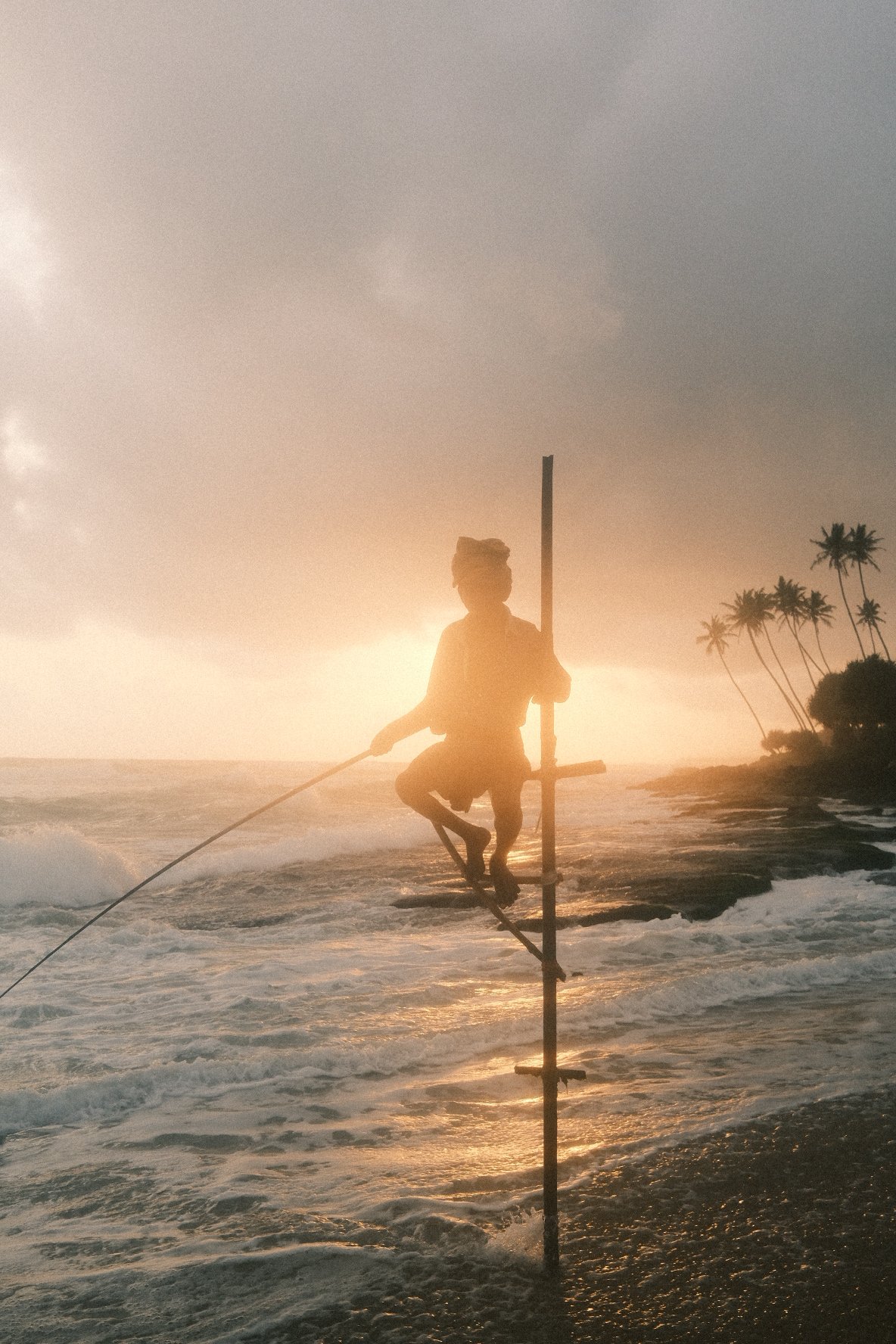 A person is practicing slacklining on the beach during sunset, with palm trees and a cloudy sky in the background.
