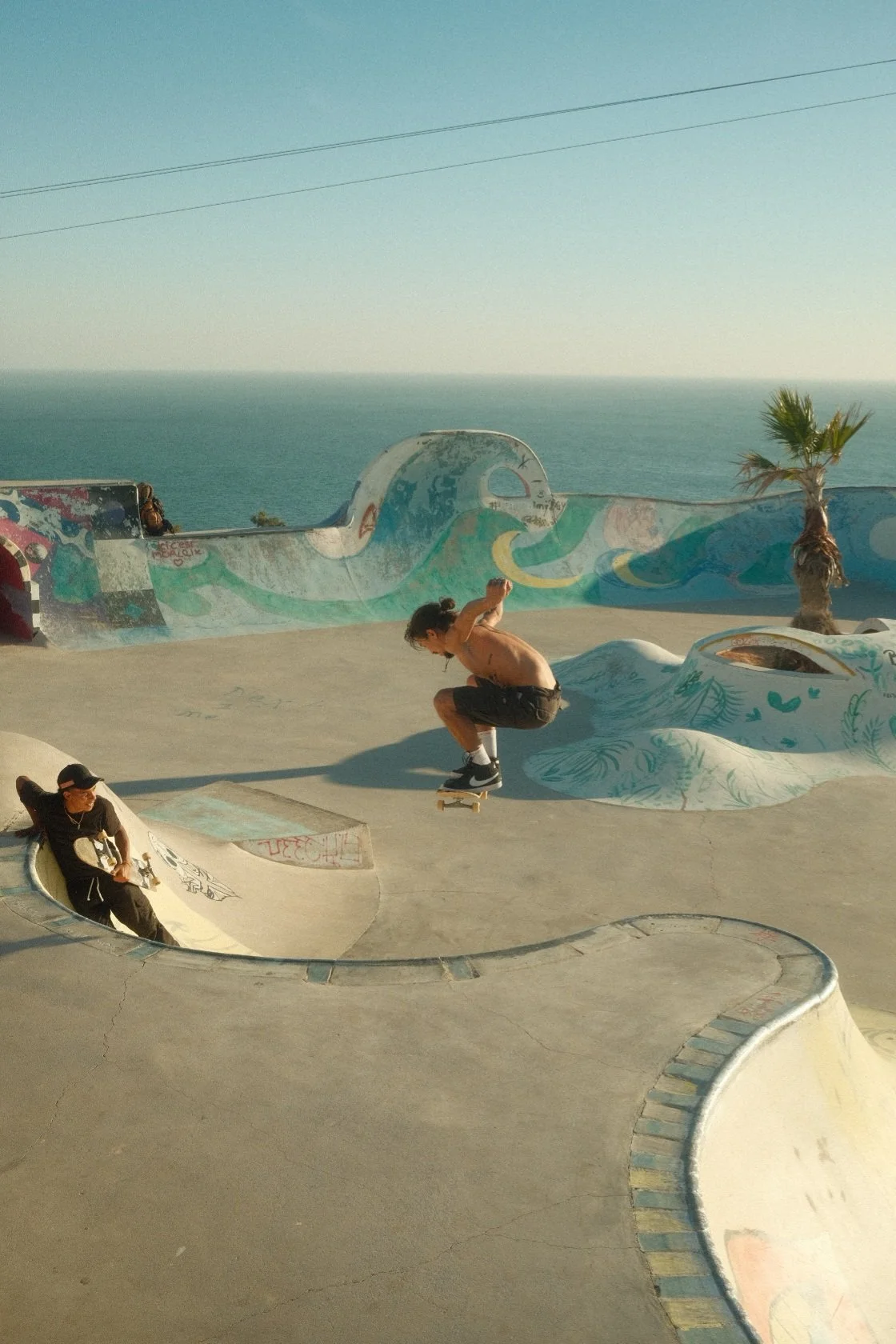 Skateboarder performing an ollie at a skateboard park near the ocean, with a palm tree and colorful murals on the ramps in Morocco.