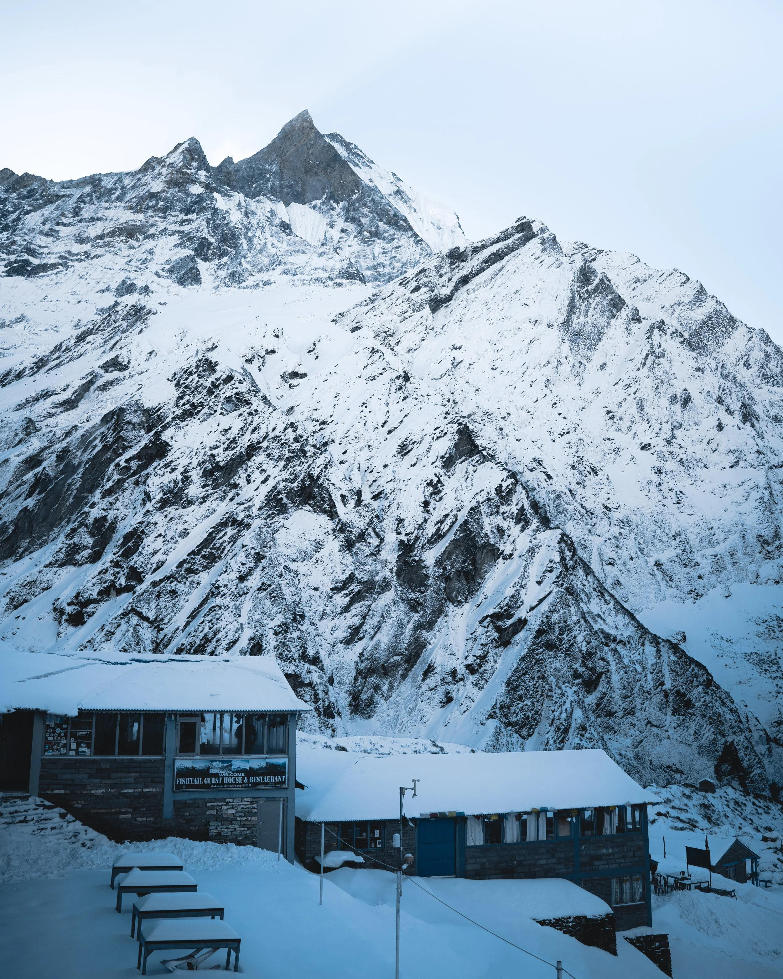 Snow-covered mountain with sharp peaks in the background and a small building labeled 'Fishtail Guest House & Restaurant' in the foreground.