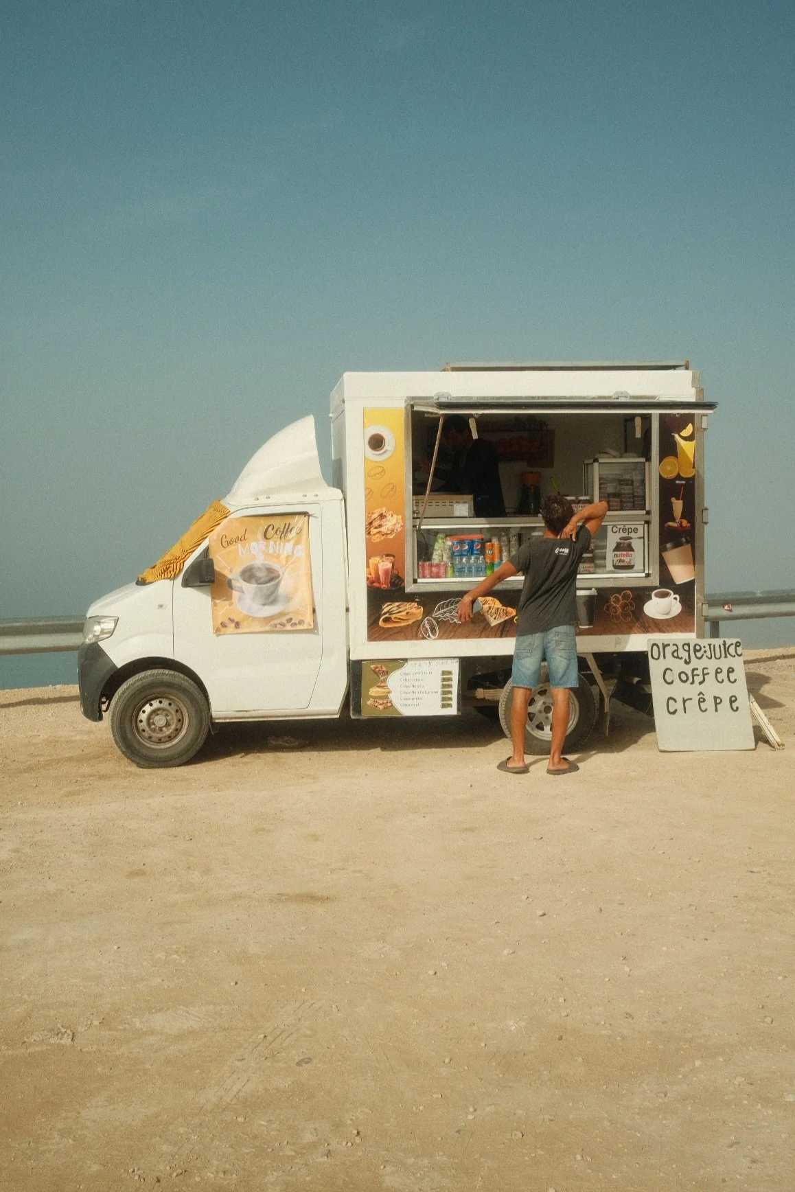 A white food truck serving orange juice, coffee, and crepes, parked on a sandy area. A person in a black t-shirt and shorts is standing in front of the truck, ordering, while the vendor is inside the truck preparing the items. The truck has colorful 
