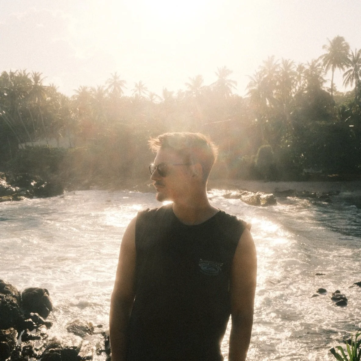 A young man with sunglasses and a sleeveless black shirt stands on rocks near the water with palm trees and the sun in the background.