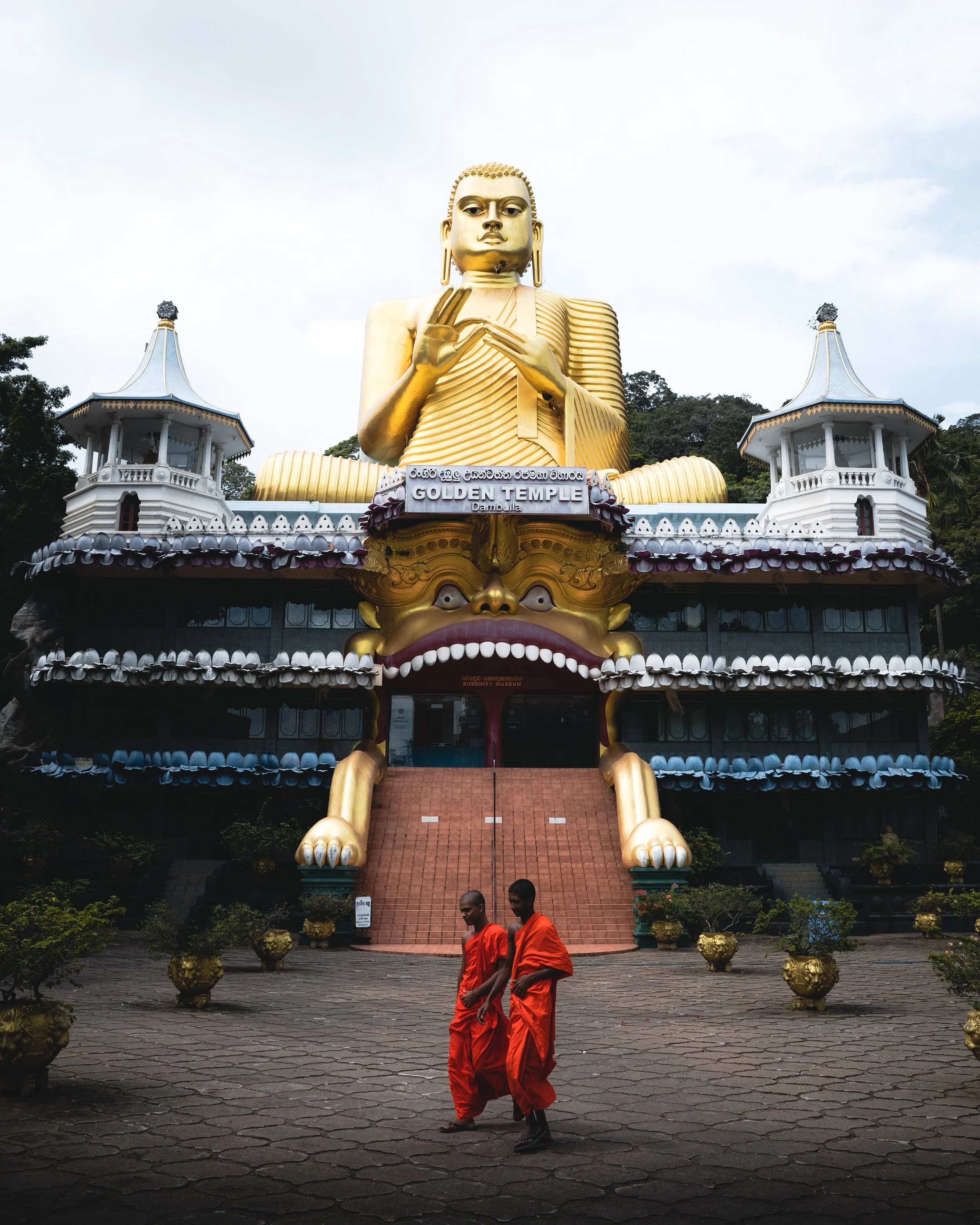 Large golden Buddha statue with a peaceful expression and hand gesture, at the entrance of a Buddhist temple, with two monks in orange robes walking in front.
