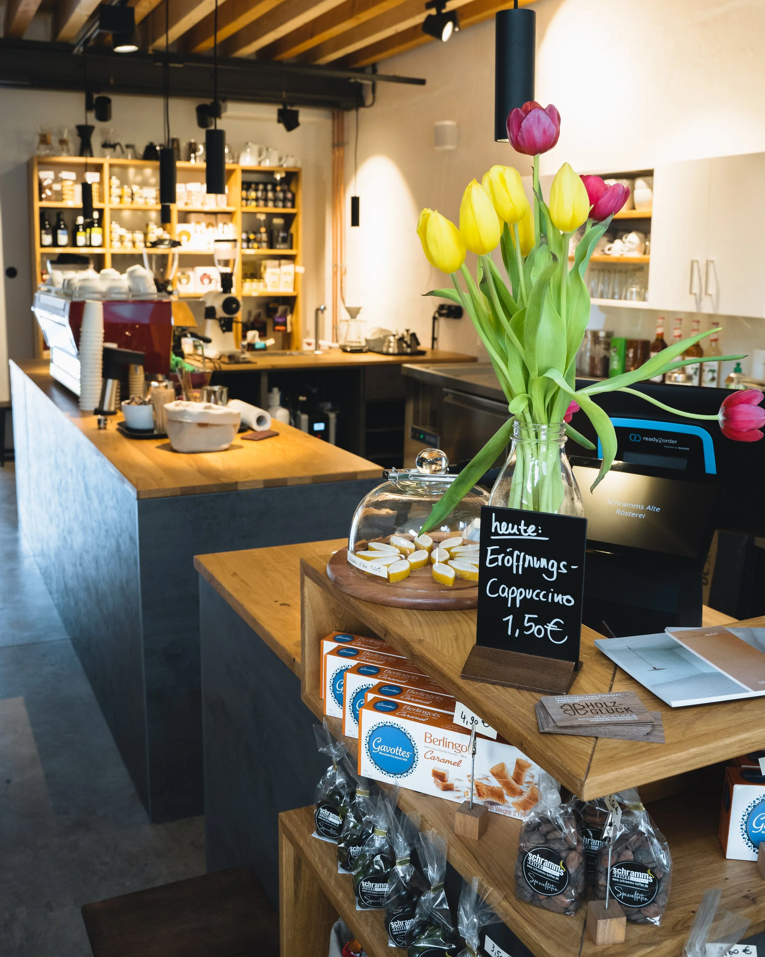 A cozy coffee shop interior with a bouquet of yellow and purple tulips in a glass vase on a wooden counter. Behind, there is a coffee machine, shelves filled with jars and cups, and a small chalkboard sign advertising cappuccino for €1.50.