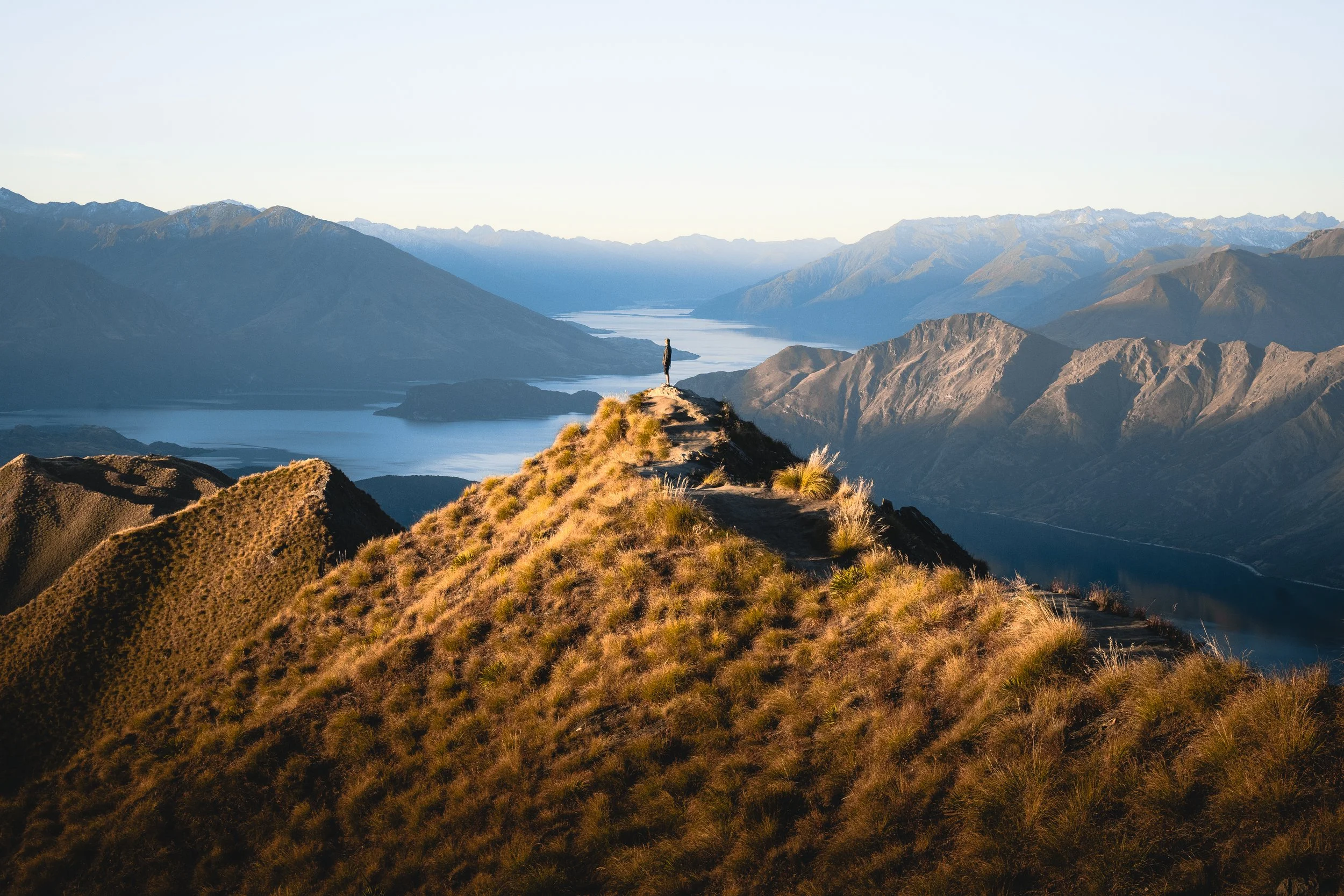 Person standing on a mountain ridge overlooking a lake and mountain range during daylight.