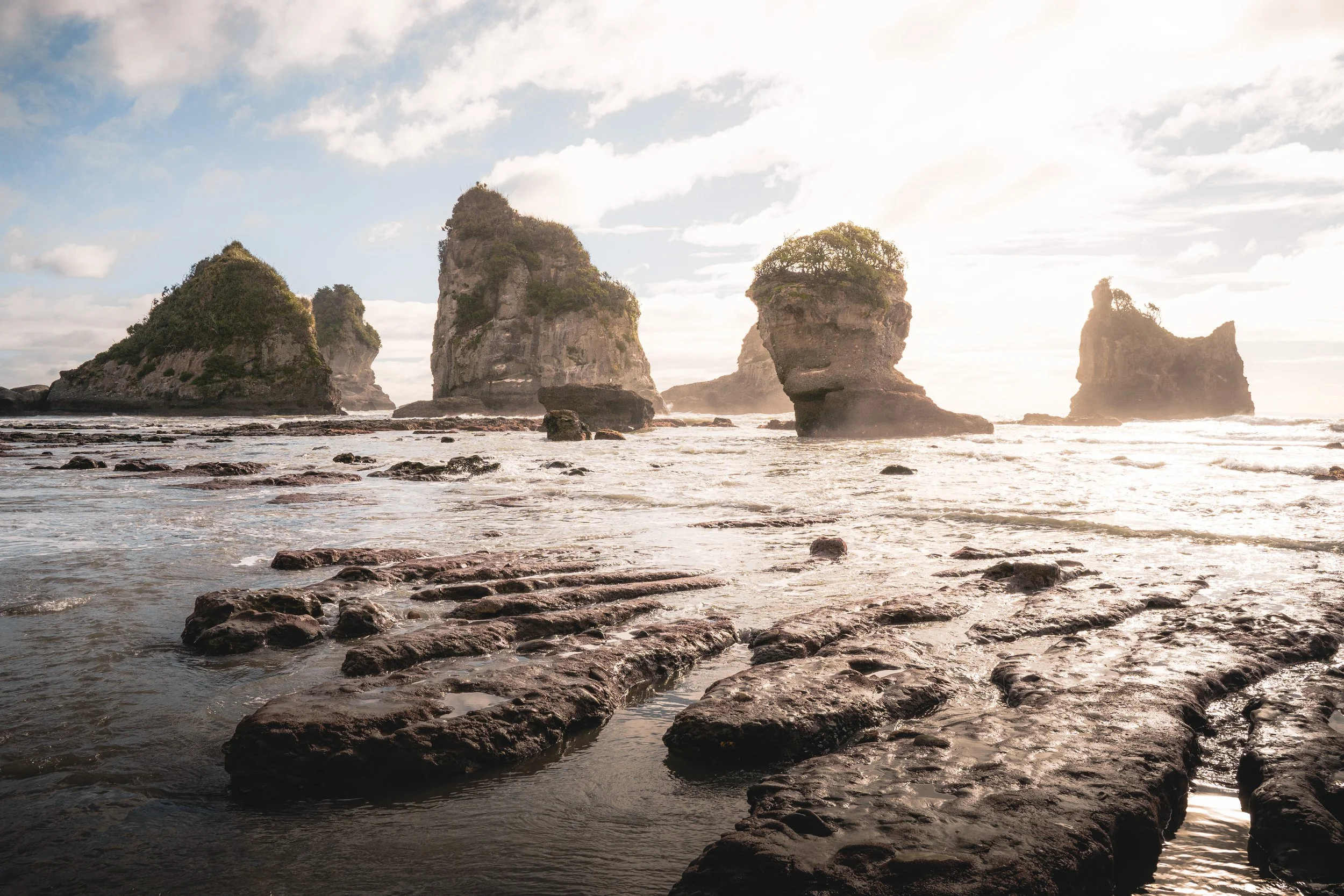 Coastal scene featuring large sea stacks with greenery on top, rocks in water, and a partly cloudy sky.