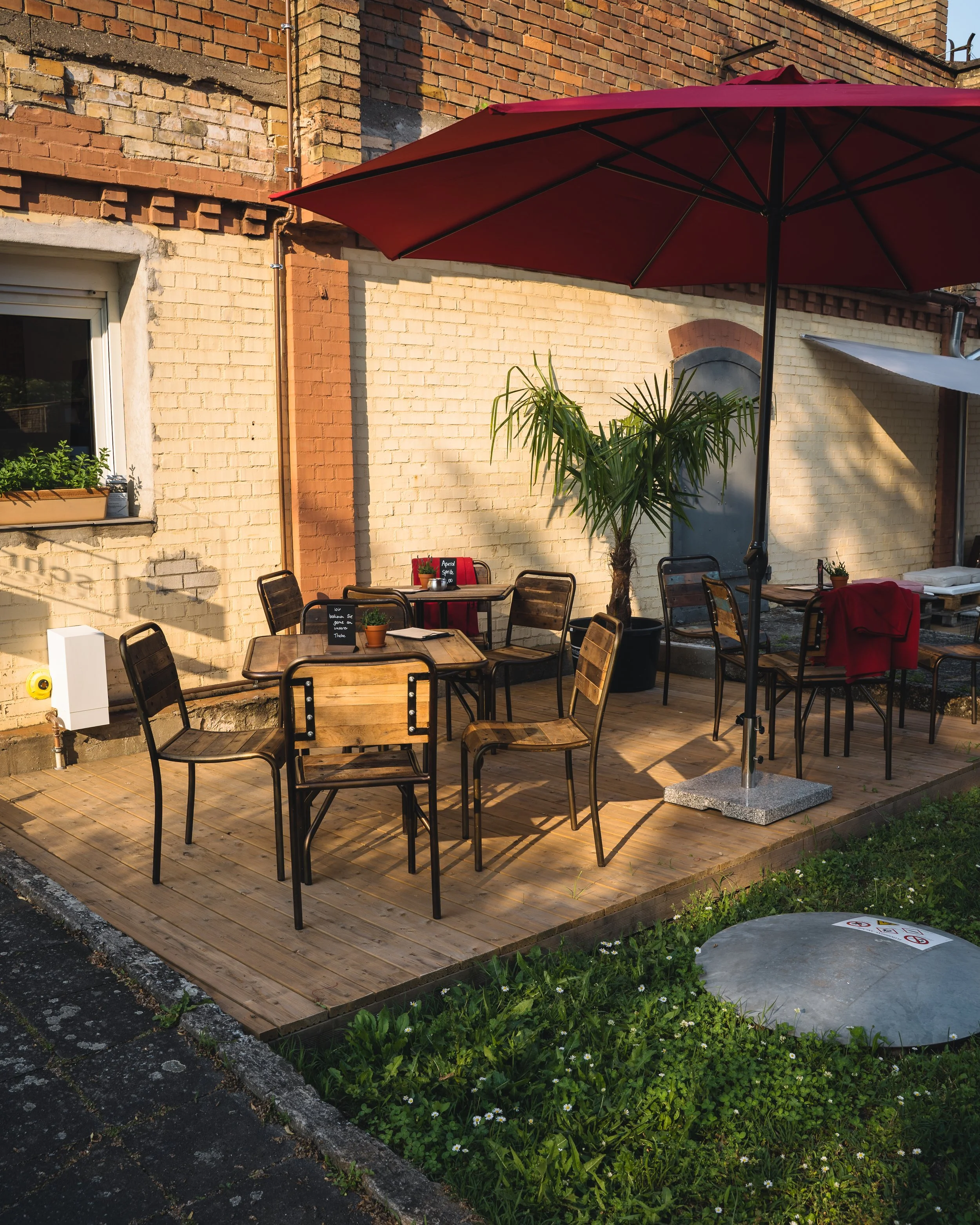 Outdoor brick wall with a large red patio umbrella providing shade over a wooden deck with tables and chairs. There are potted plants and a small flower bed with green plants and white flowers in the foreground.