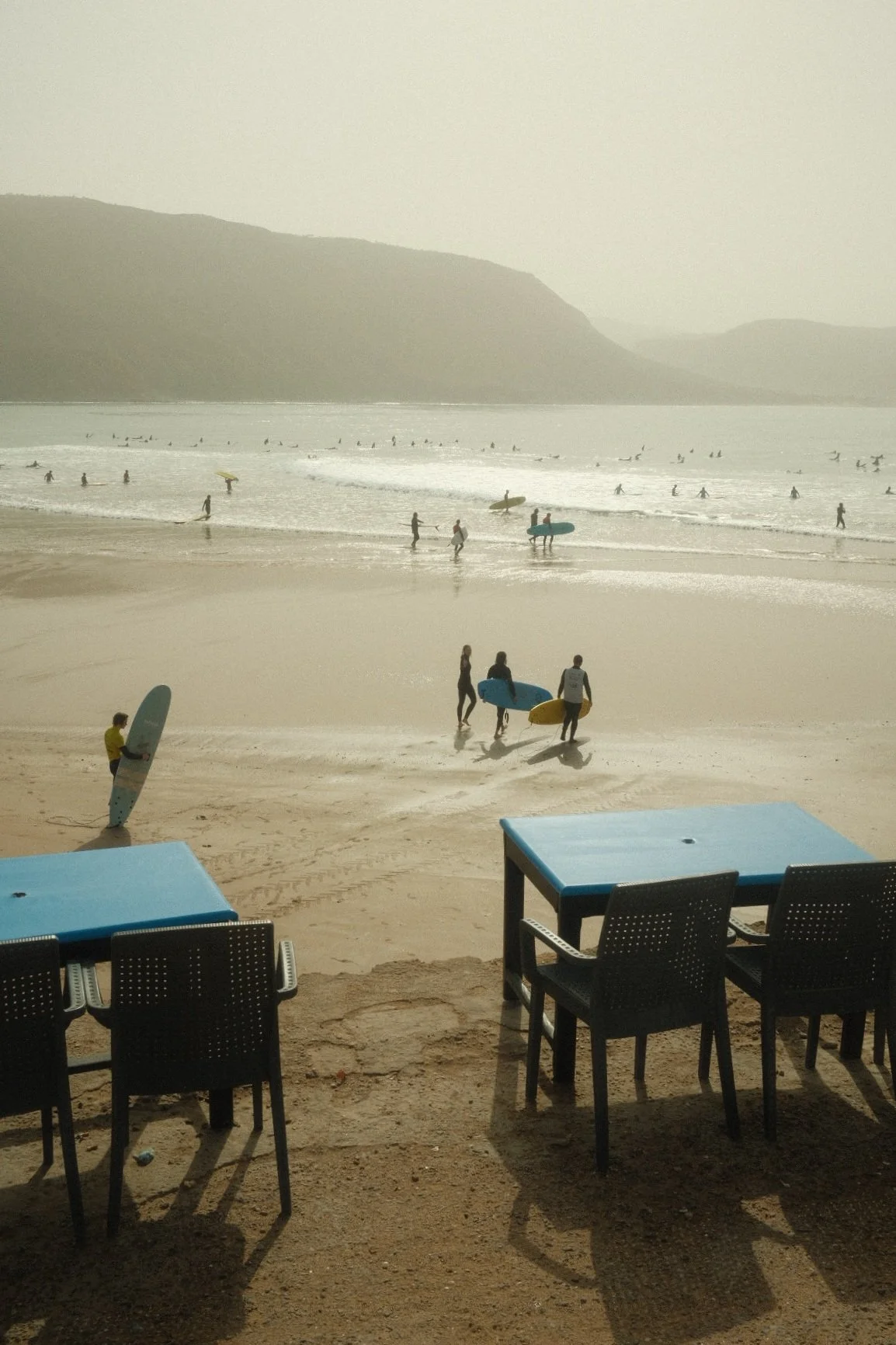 People at the beach with surfboards, some walking towards the water, others surfing, and chairs and tables in the foreground.