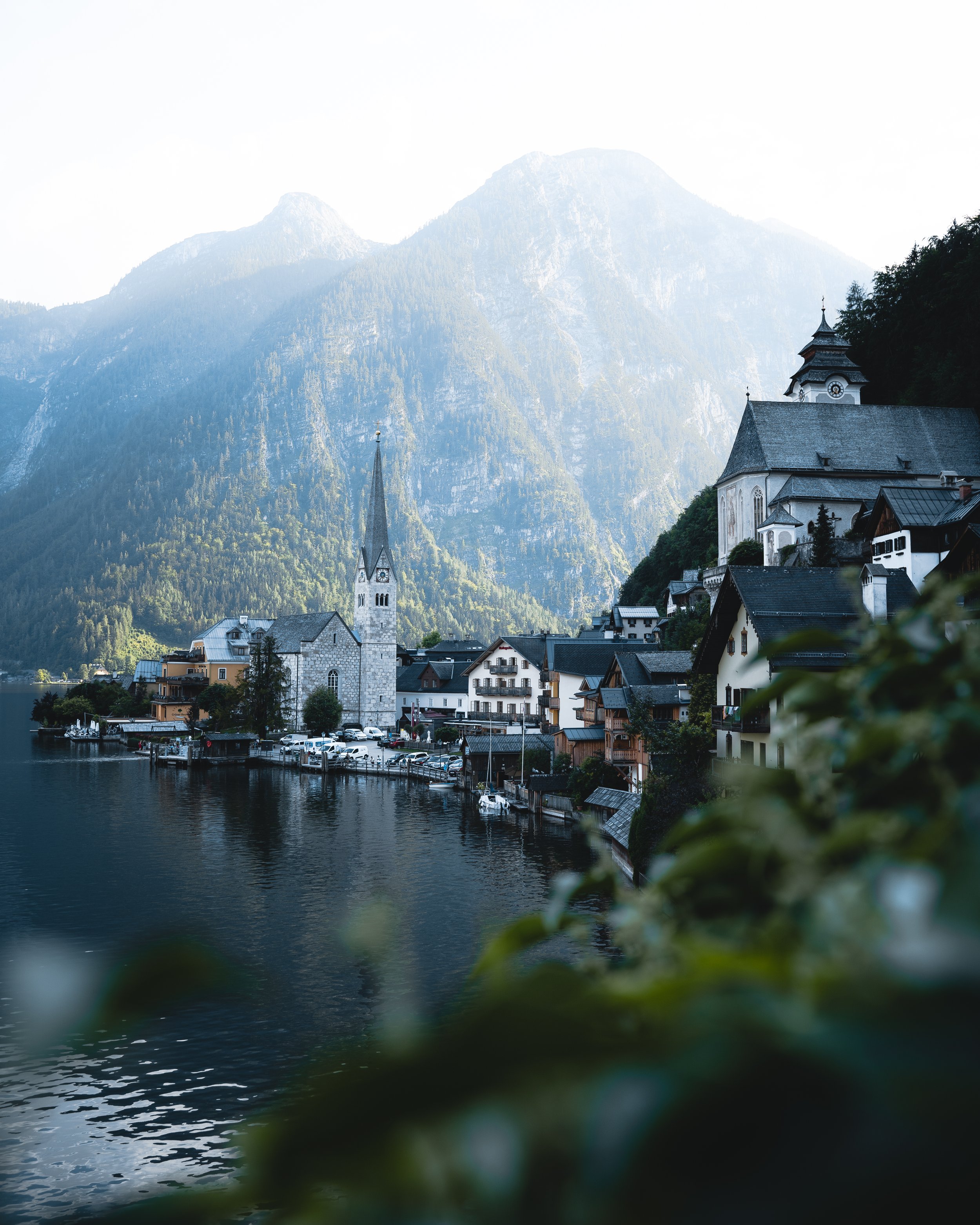 A serene lakeside village with church steeples, surrounded by lush green mountains and a calm body of water in the foreground.