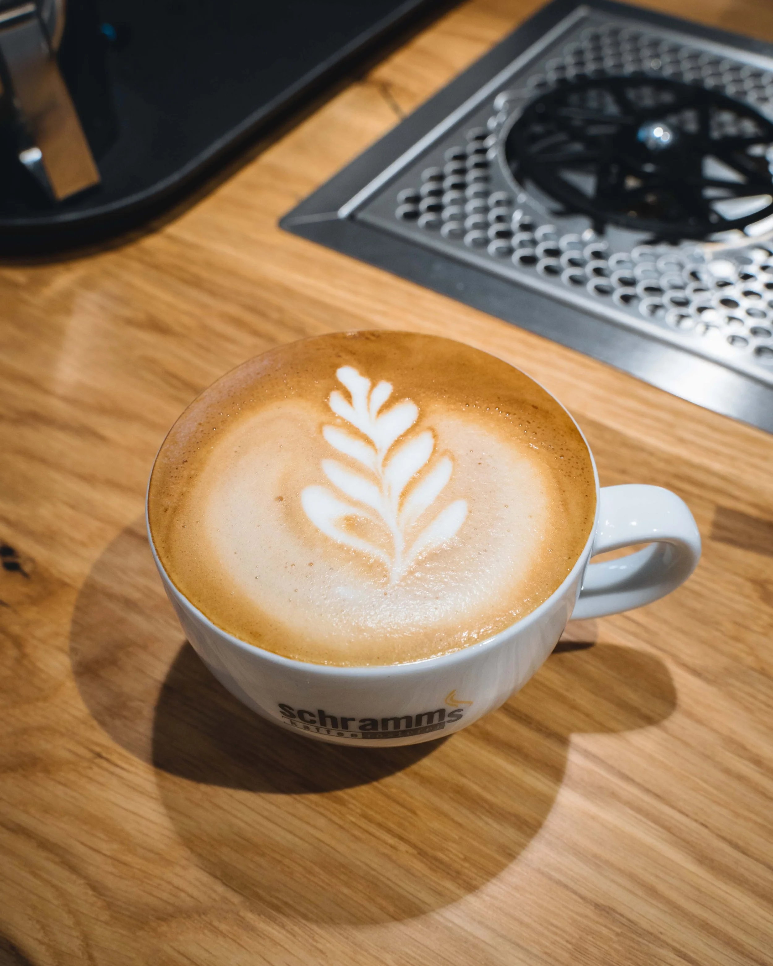 Close-up of a white ceramic mug with latte art on top, sitting on a wooden table. The latte art is in the shape of a leaf. In the background, part of a computer keyboard and a black electronic device are visible.