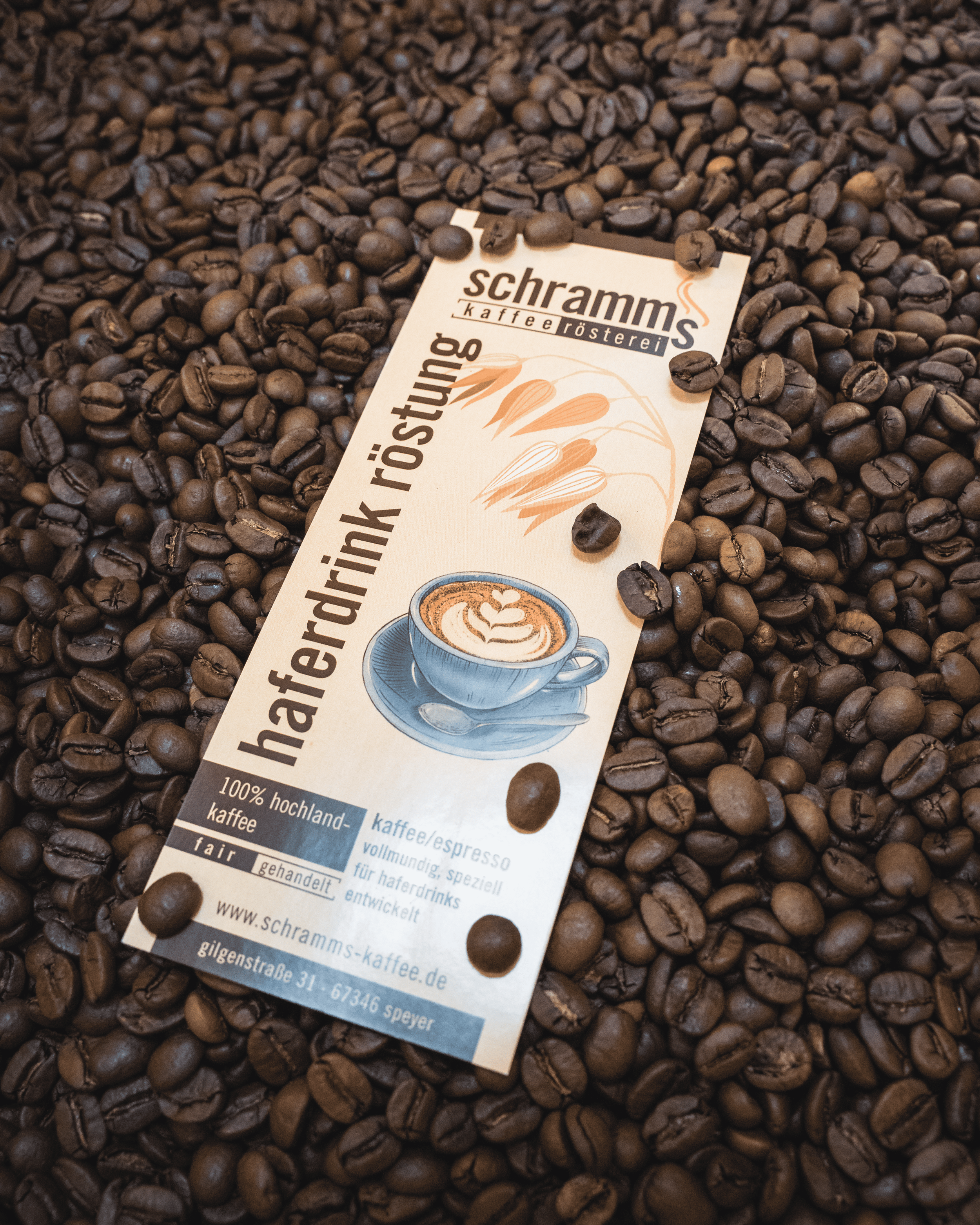 Close-up of roasted coffee beans with a partially visible coffee shop business card lying on top. The business card includes a coffee cup illustration and text in German.