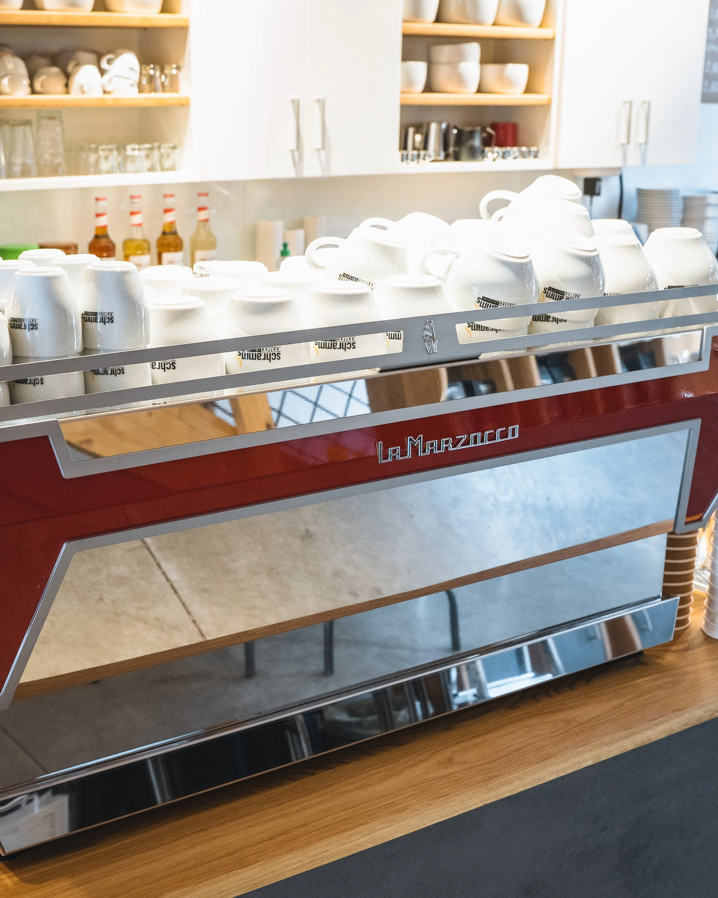 Coffee mugs with a 'Schrams' logo on a metal rack, set in a cafe or restaurant kitchen.