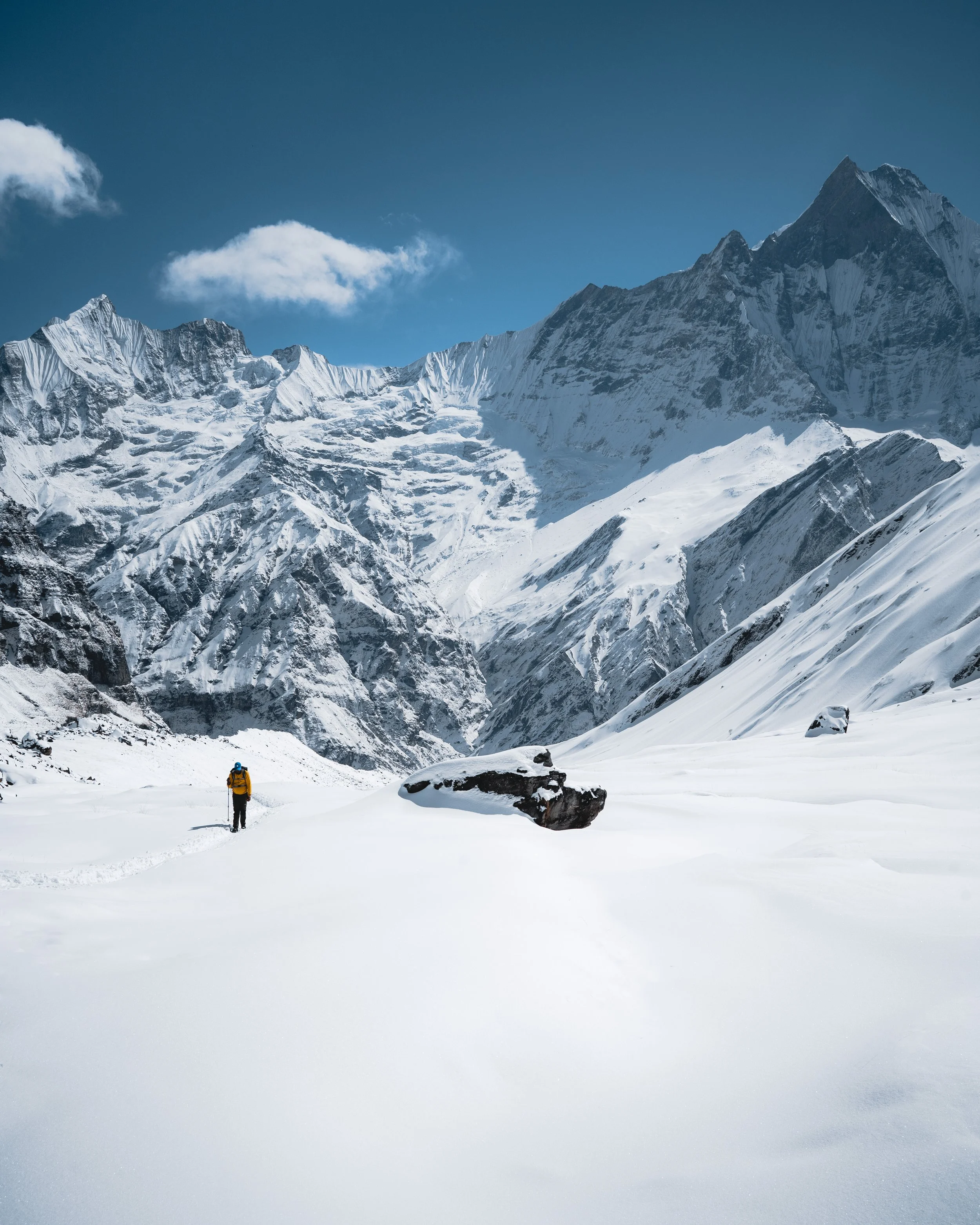 A lone hiker dressed in a yellow jacket, black pants, and carrying a backpack stands in a vast snow-covered mountain landscape, with towering, snow-capped peaks under a clear blue sky with a few clouds.