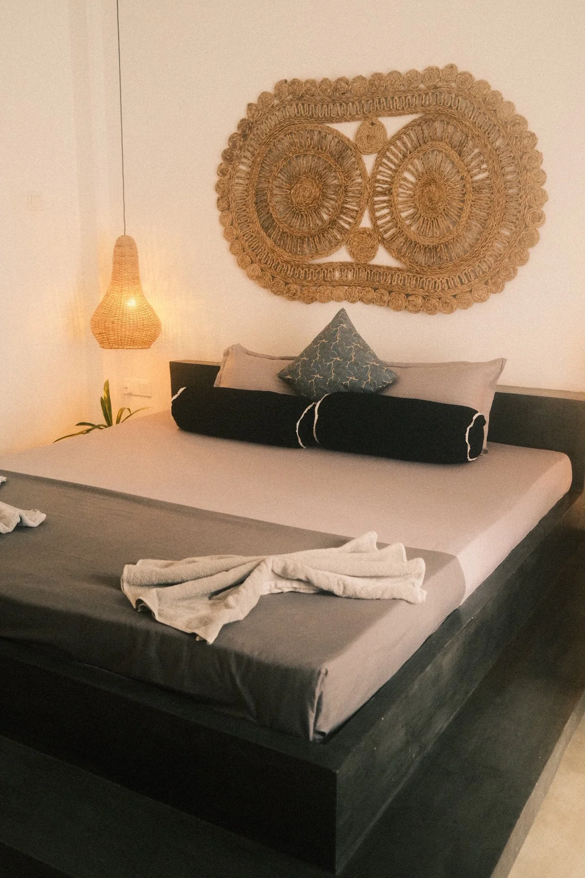 A bedroom with a bed, gray sheets, and various pillows, including a triangular patterned pillow. A woven wall art piece is above the bed, and a hanging woven lamp is on the side. A towel or piece of clothing is on the bed.