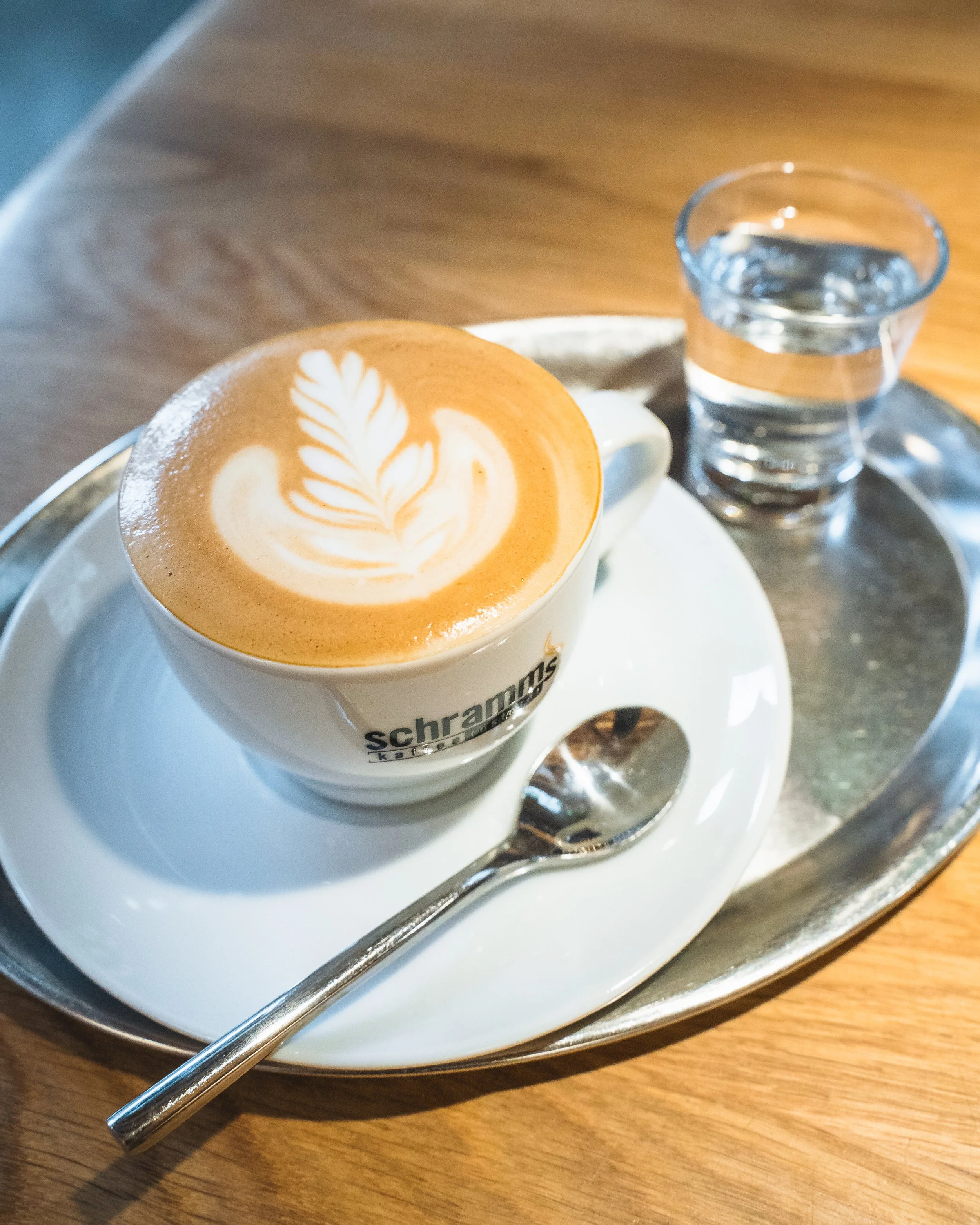 A white ceramic cup with latte art on top, placed on a white saucer on a metallic tray with a silver straw and a small glass of water beside it on a wooden table.