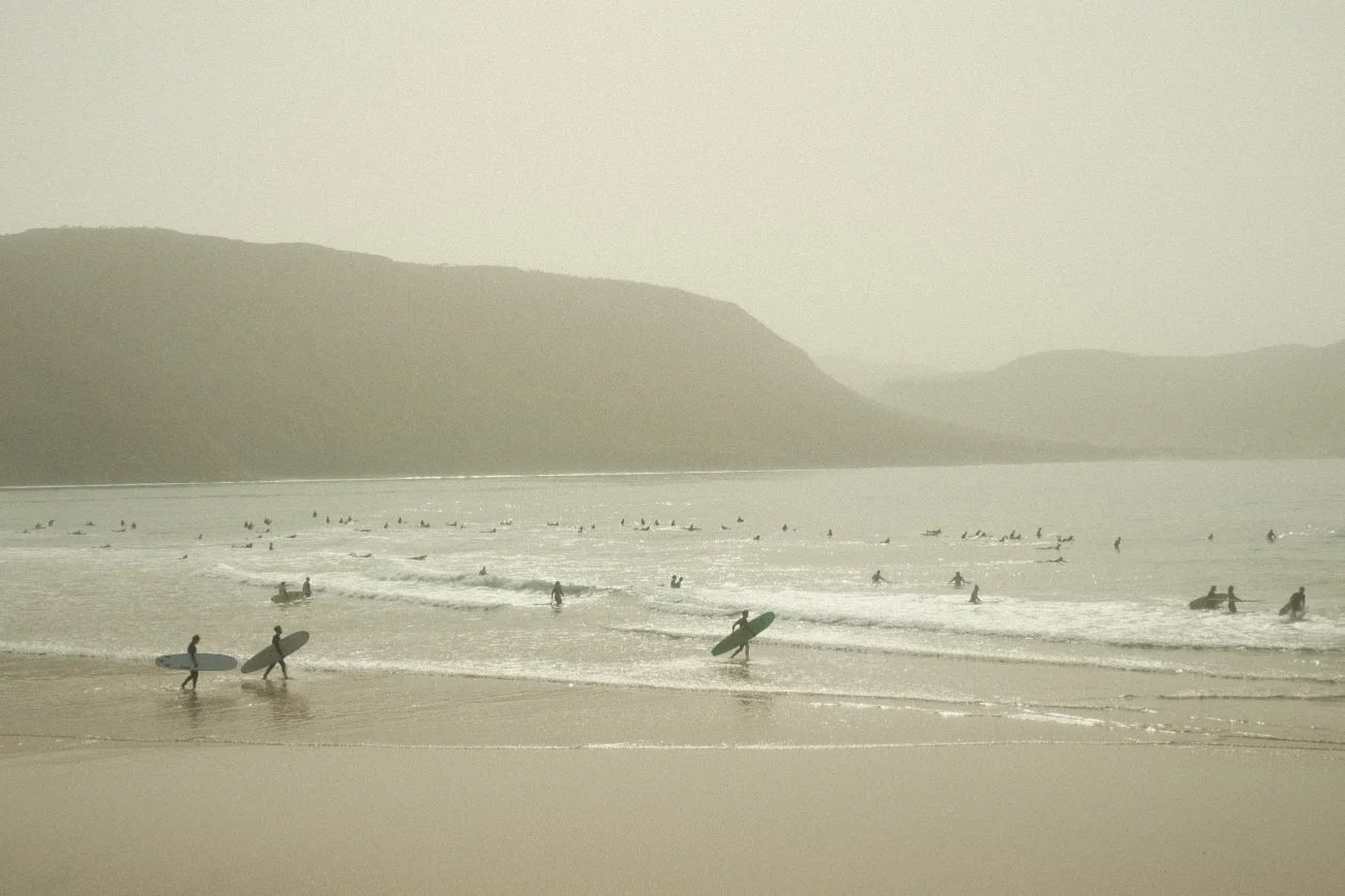 People surfing and walking on the beach with surfboards, with a mountain in the background on a cloudy day.