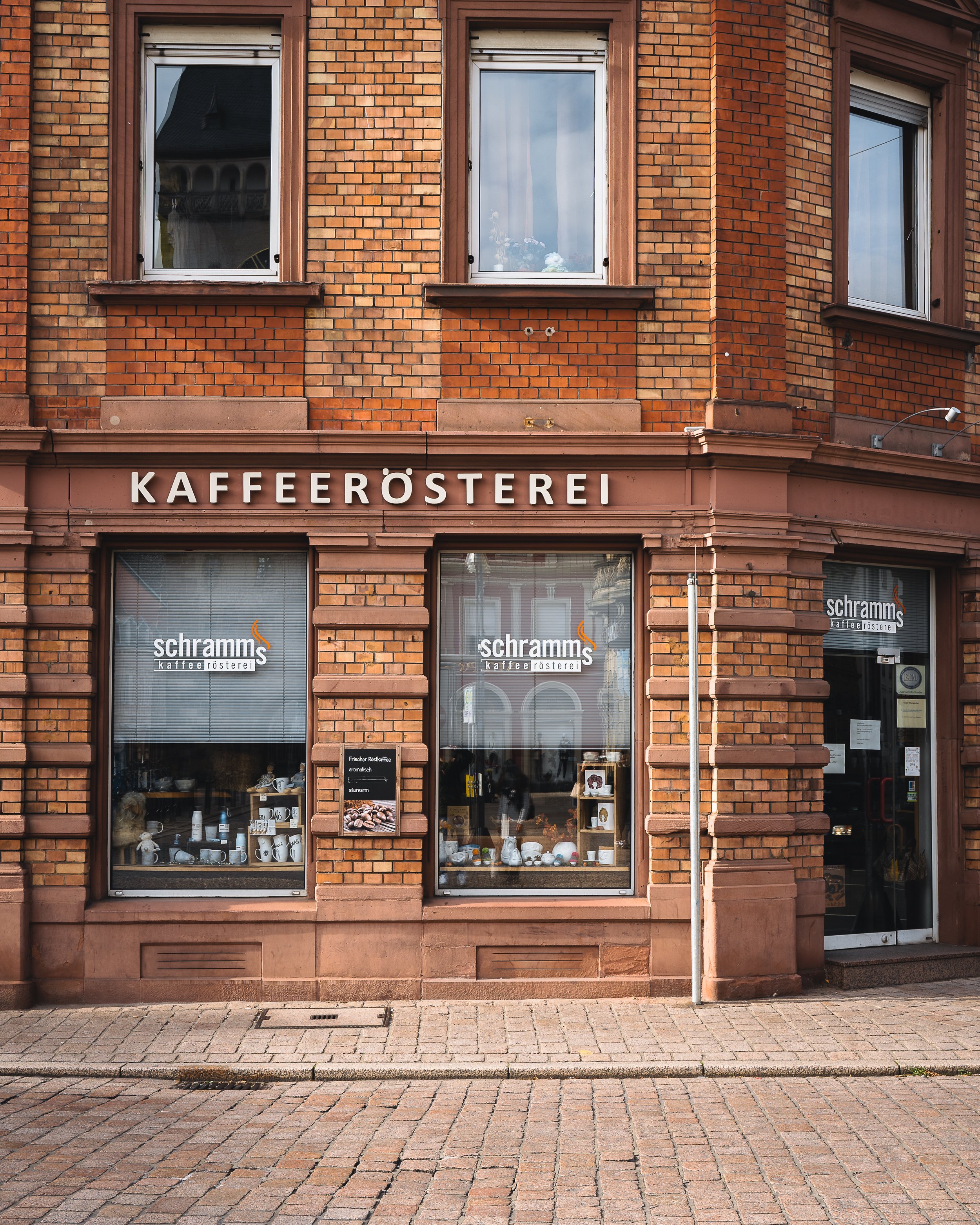 The image shows the front of a brick building with three large windows and a sign that reads "KAFFERÖSTEREI" above the door. The windows display various items, including white mugs and decorative objects. The sidewalk in front is made of stone, and t