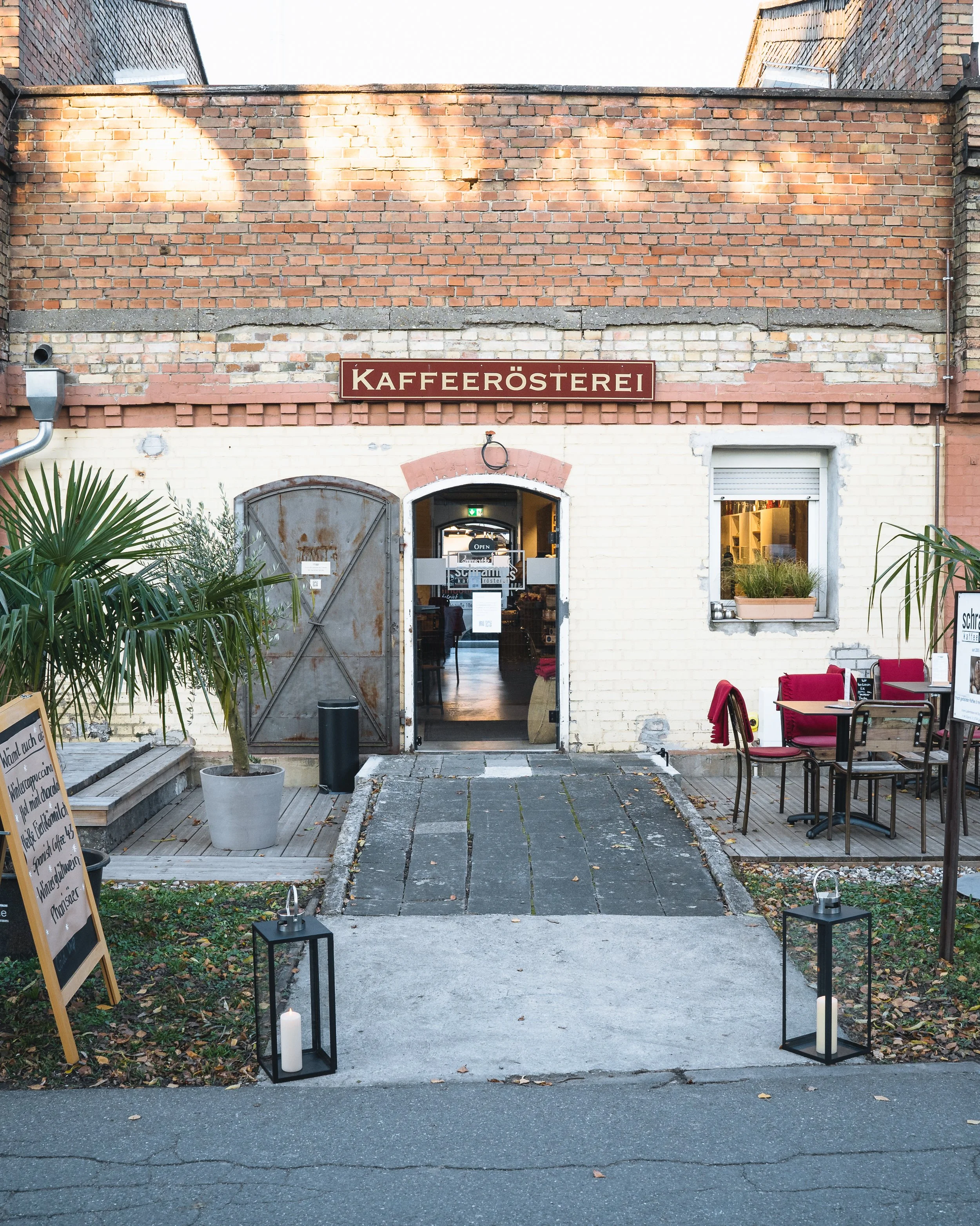 Exterior view of a coffee shop with a sign reading 'KAFFERÖSTEREI' on a brick wall. Outdoor seating area with tables, chairs, and candlelit lanterns, leading to the shop's entrance.