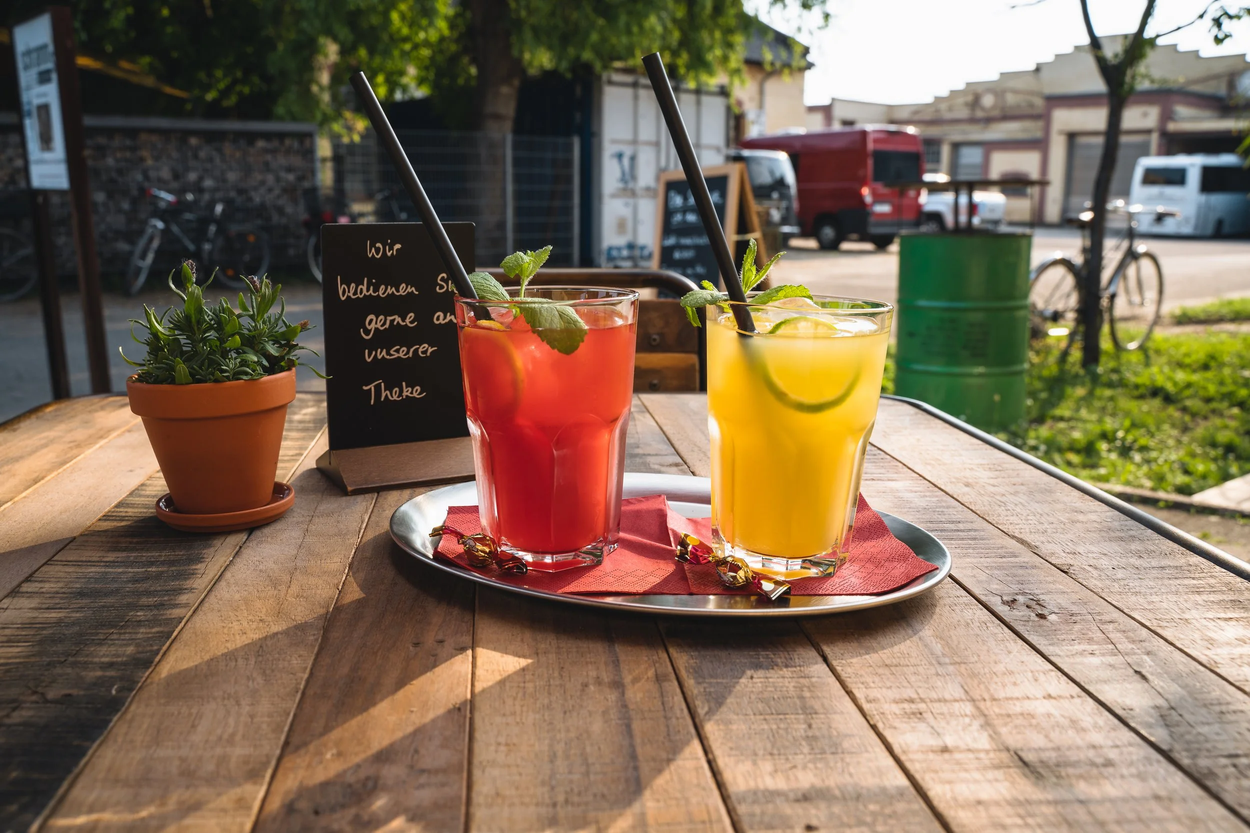 Two colorful cocktails, one red and one yellow, served on a metal tray on a wooden outdoor table. A potted plant and a small chalkboard sign are also on the table, with a city street scene in the background.