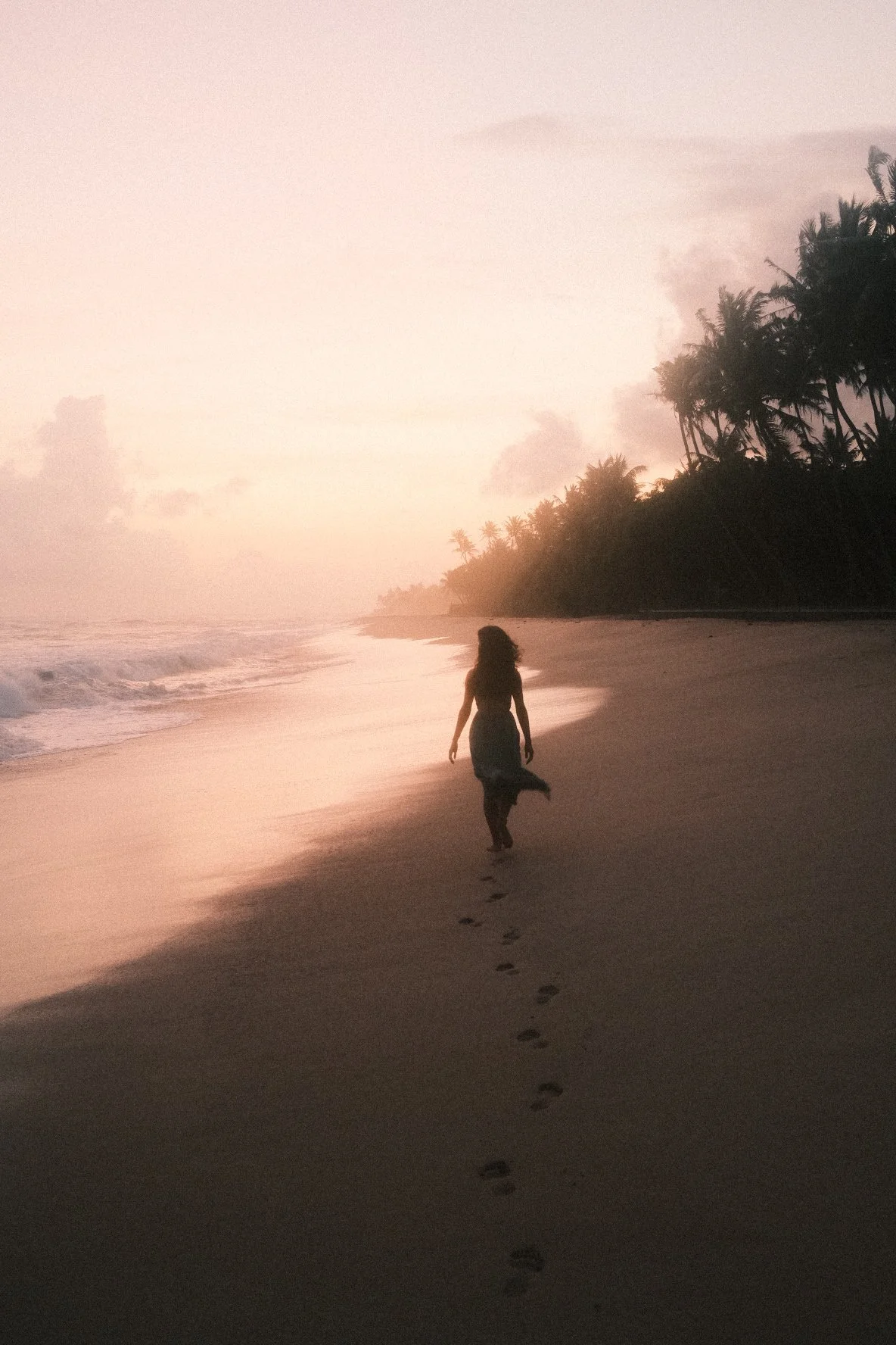 A woman walking along a beach at sunset with palm trees in the background and footprints in the sand.