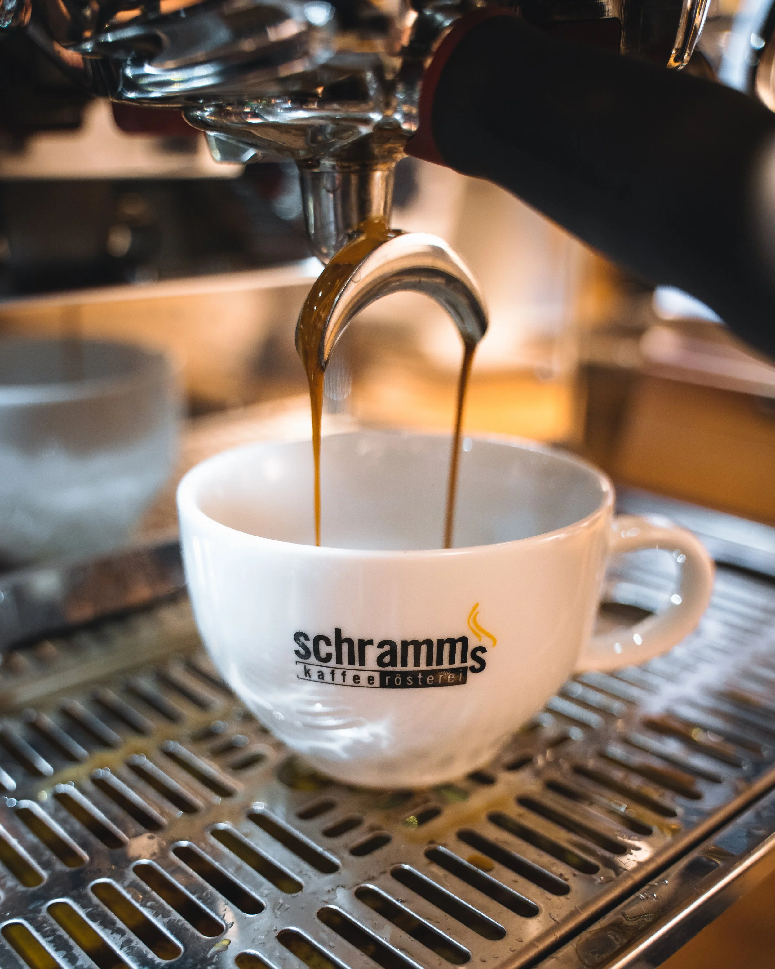 Close-up of dark coffee being brewed into a white mug on a metallic drip tray, with the mug featuring the logo 'schramm's kaffeerösterei'.